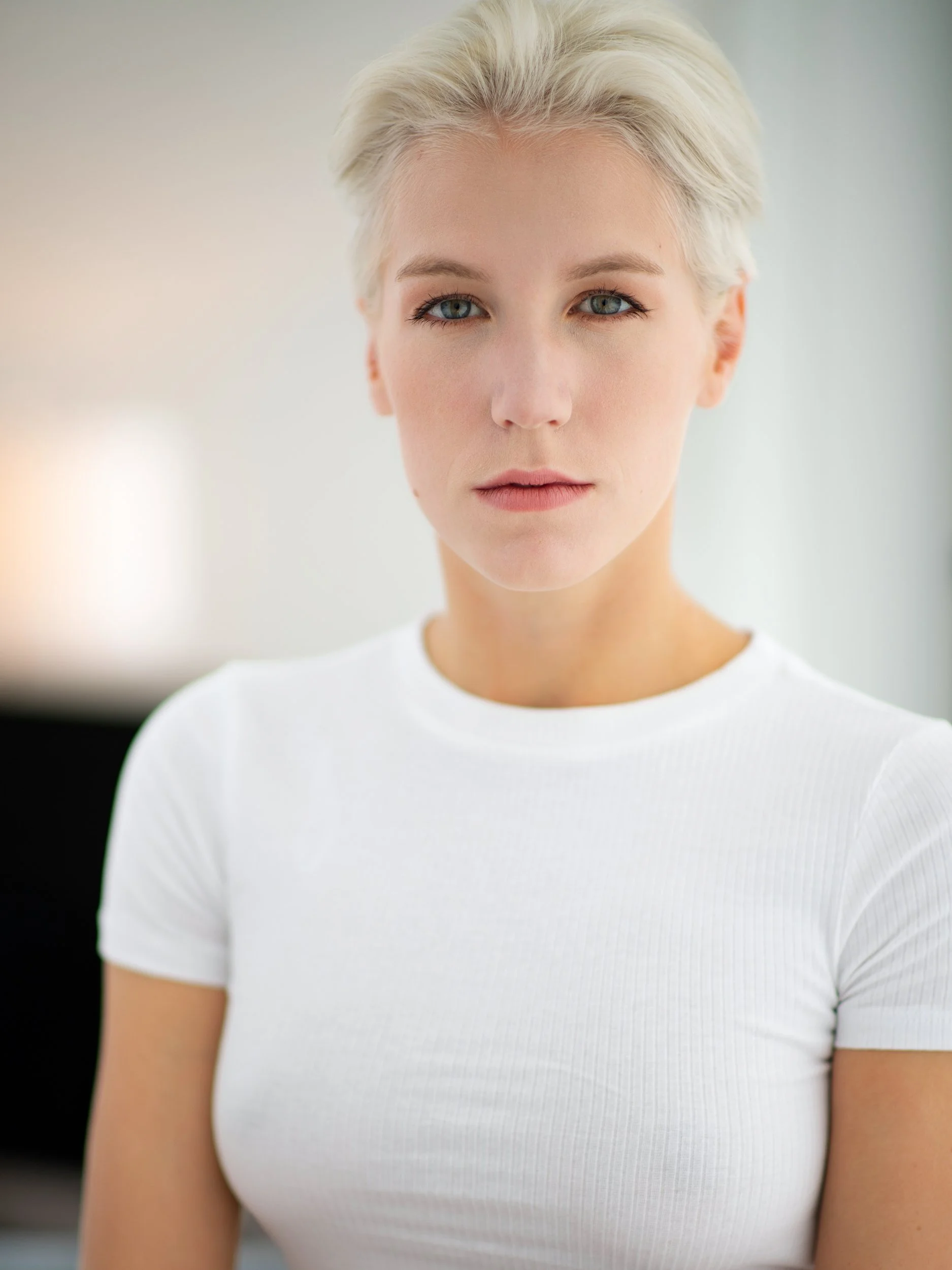 Helga with a still, direct gaze, cropped platinum-blonde hair and pale blue eyes, simple white tee, warm bokeh in soft natural light.