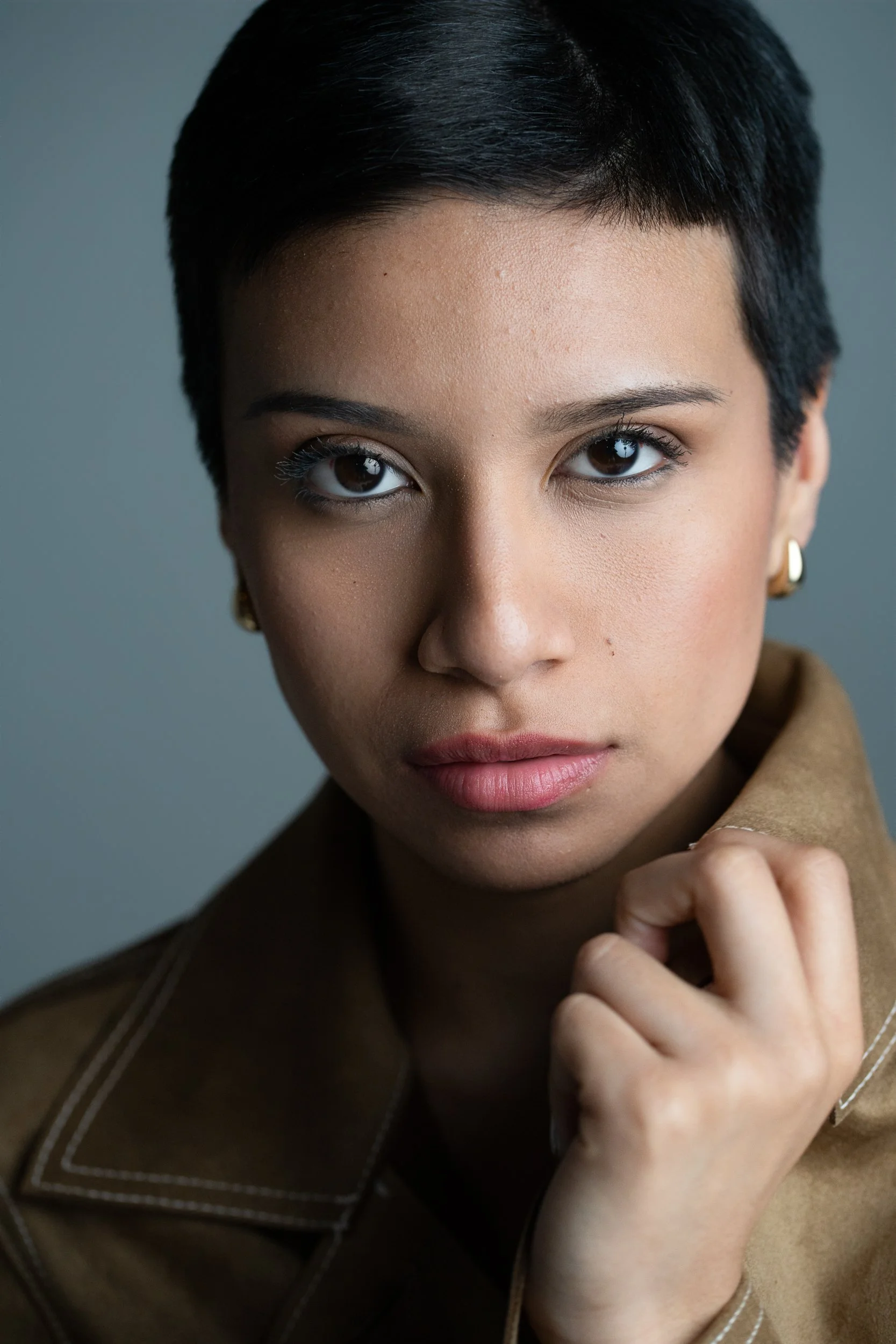 Amanda, portrait session at 9 Studios, SoHo NYC — close editorial portrait, direct gaze, tan leather jacket with contrast stitching, hand at collar, gray background, May 2026