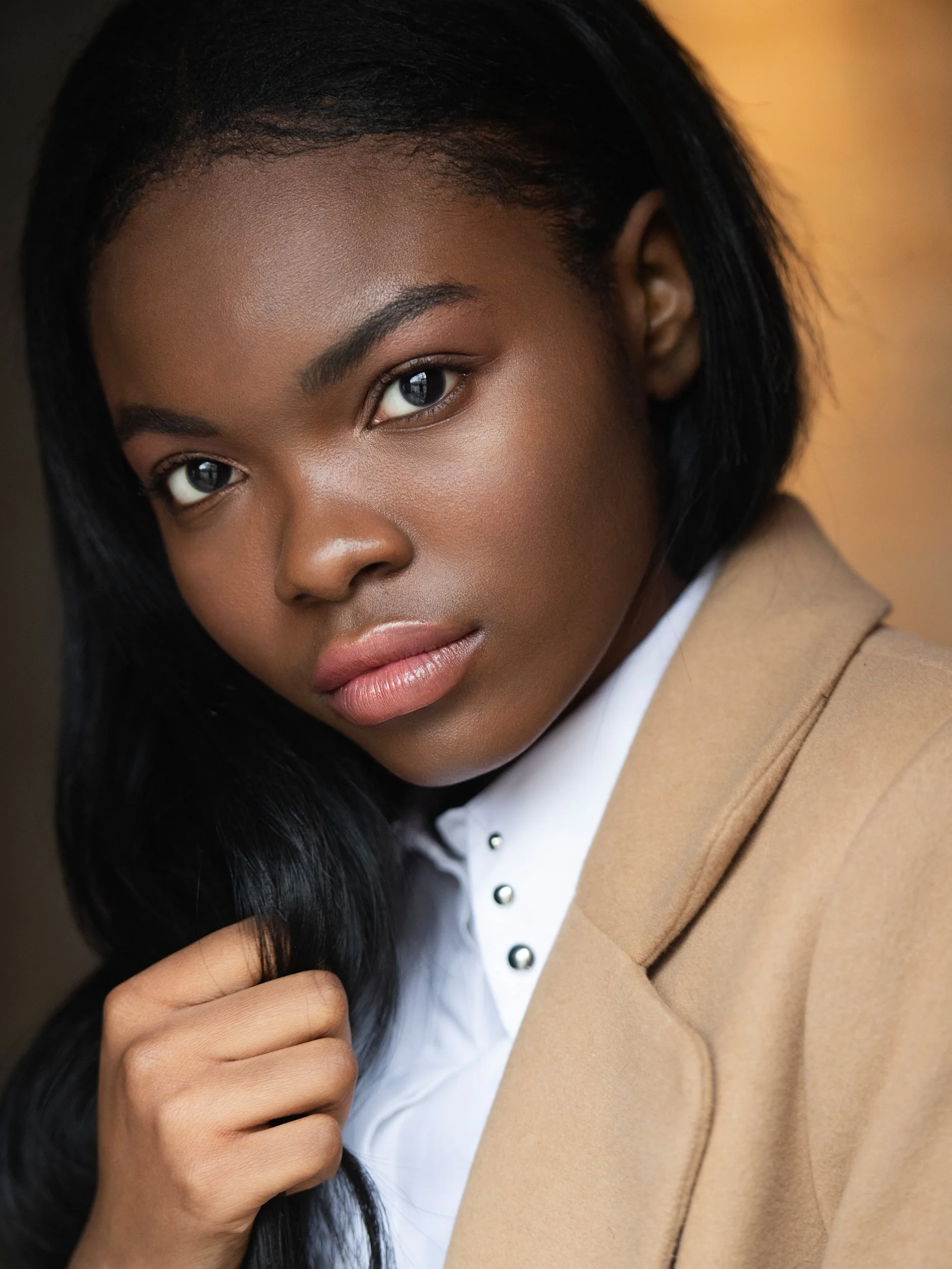 Mia in a close beauty portrait, steady direct gaze, sleek black hair and a white collared shirt under a camel coat, warm amber light.