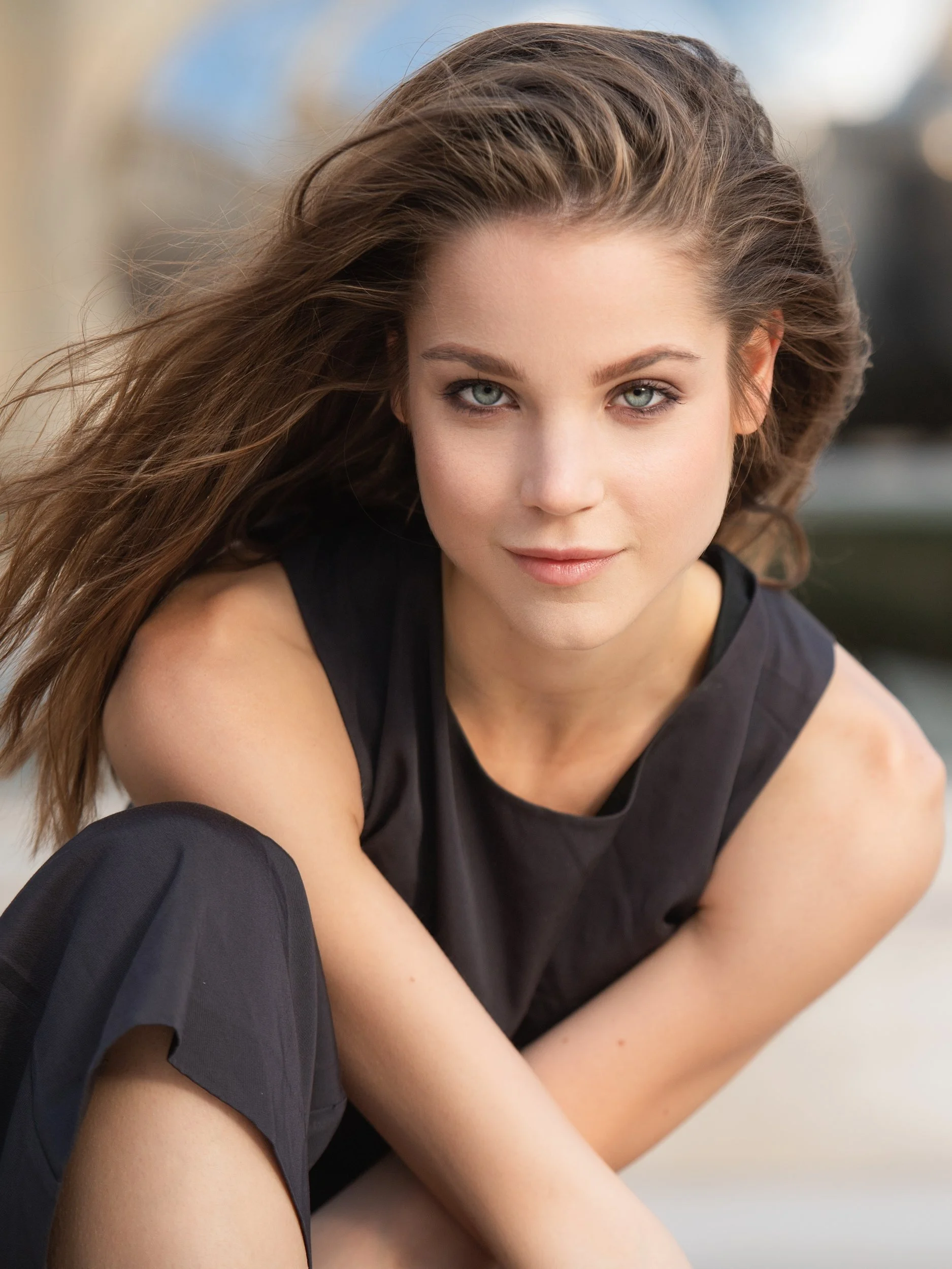 Paulina kneeling with a calm, level gaze, windblown brown hair and pale green eyes, black sleeveless top, soft sunlit bokeh on a Paris street.