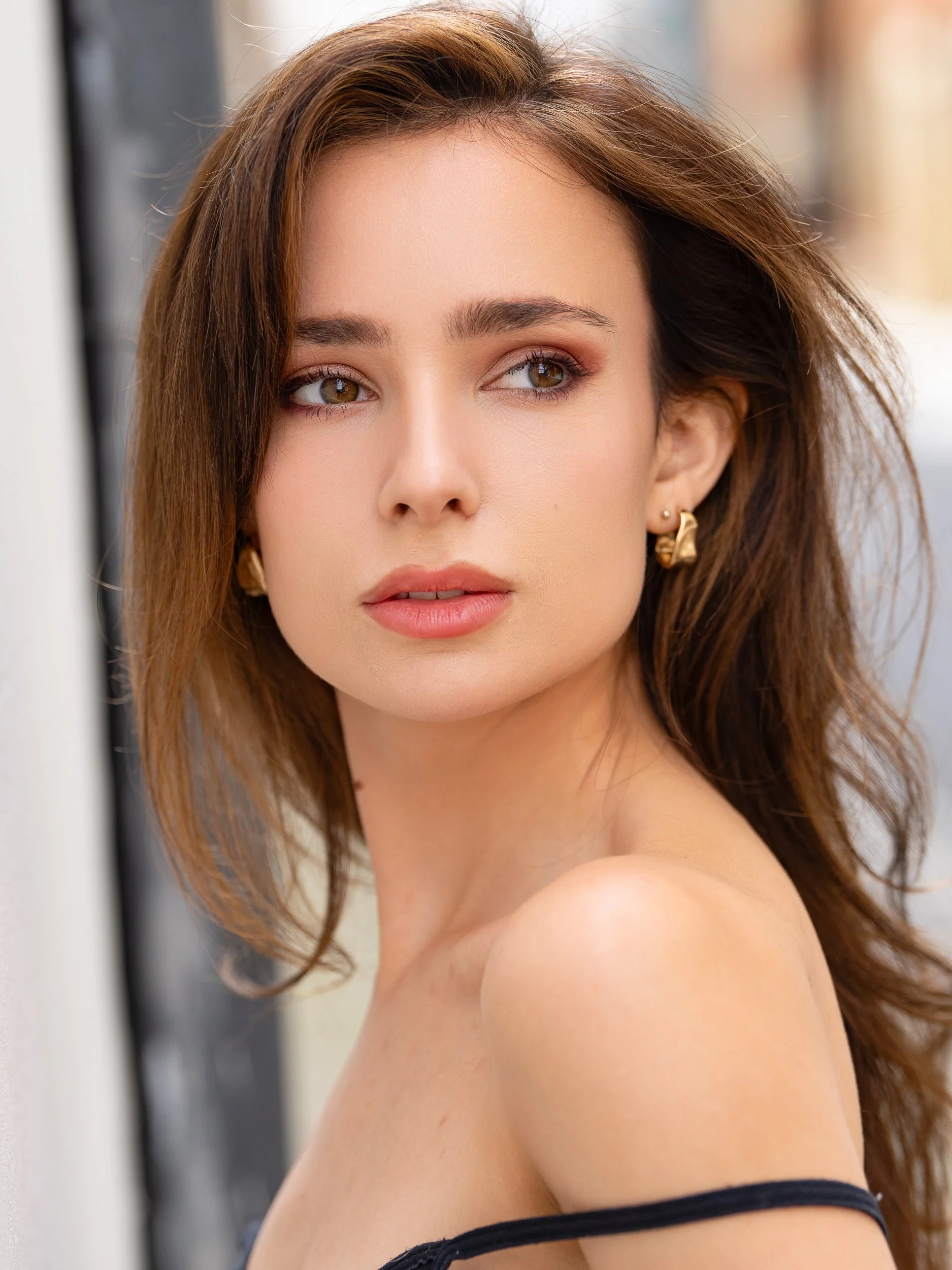 Emma with a soft, distant gaze turned over her shoulder, long brown hair and bold gold earrings, warm natural light on a blurred London street.