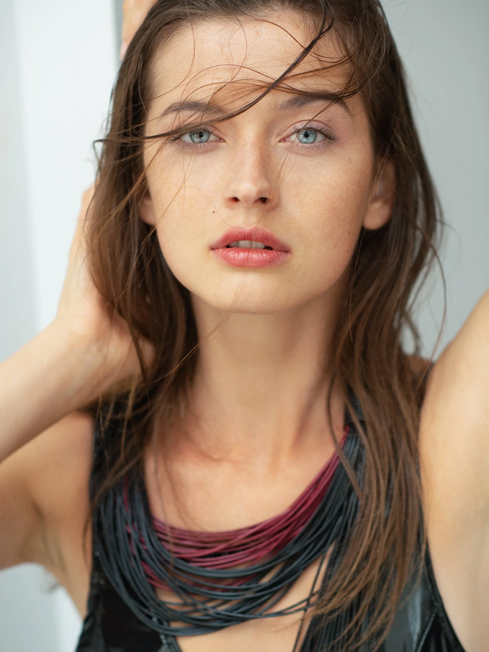 Anna gazing up with hands lifted to her damp, windblown hair, freckled skin and pale eyes, multi-strand red-and-black necklace, bright studio.