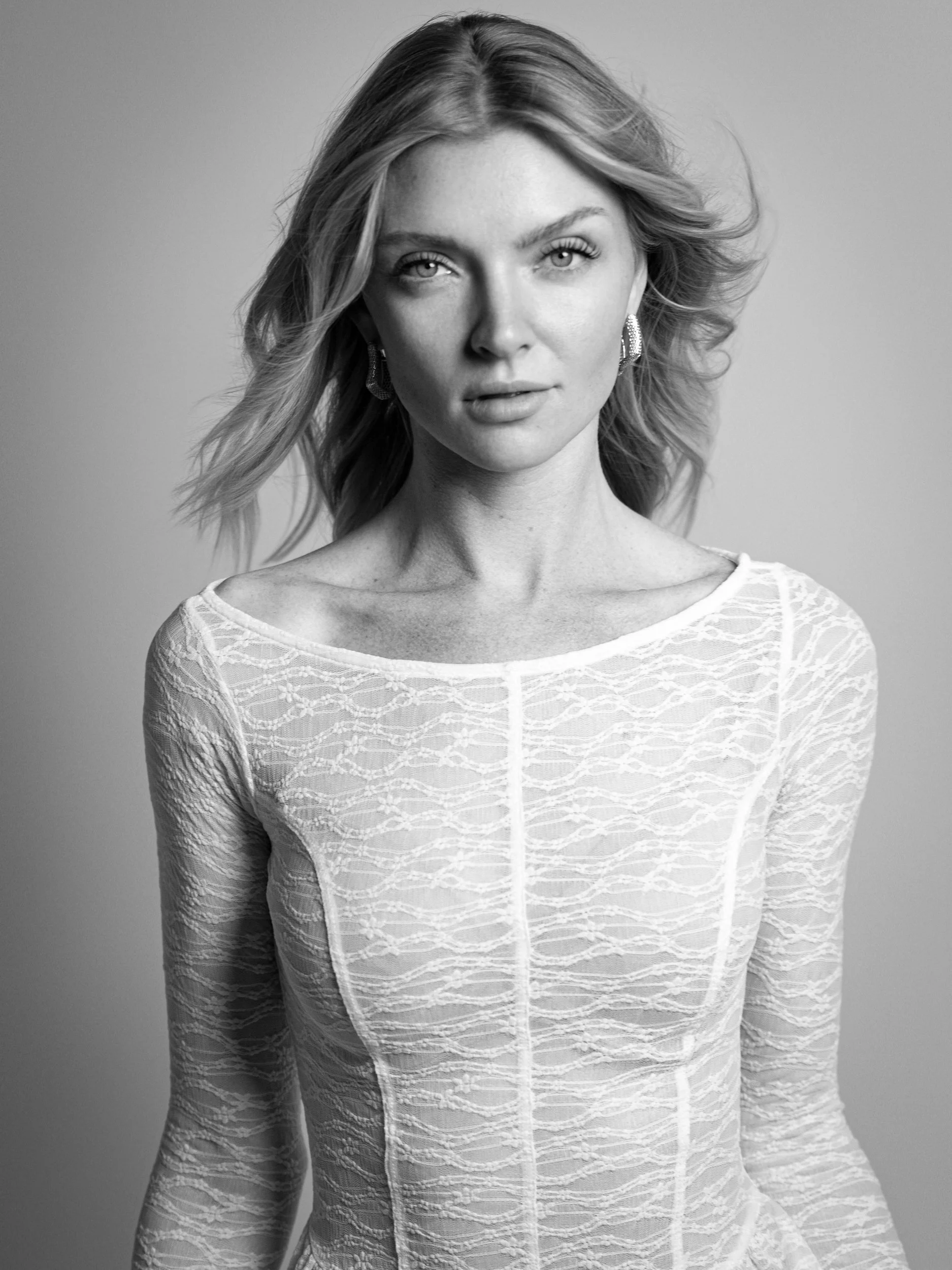 Whitney, black and white portrait in white lace dress with direct gaze, windswept blonde hair, statement earrings, studio