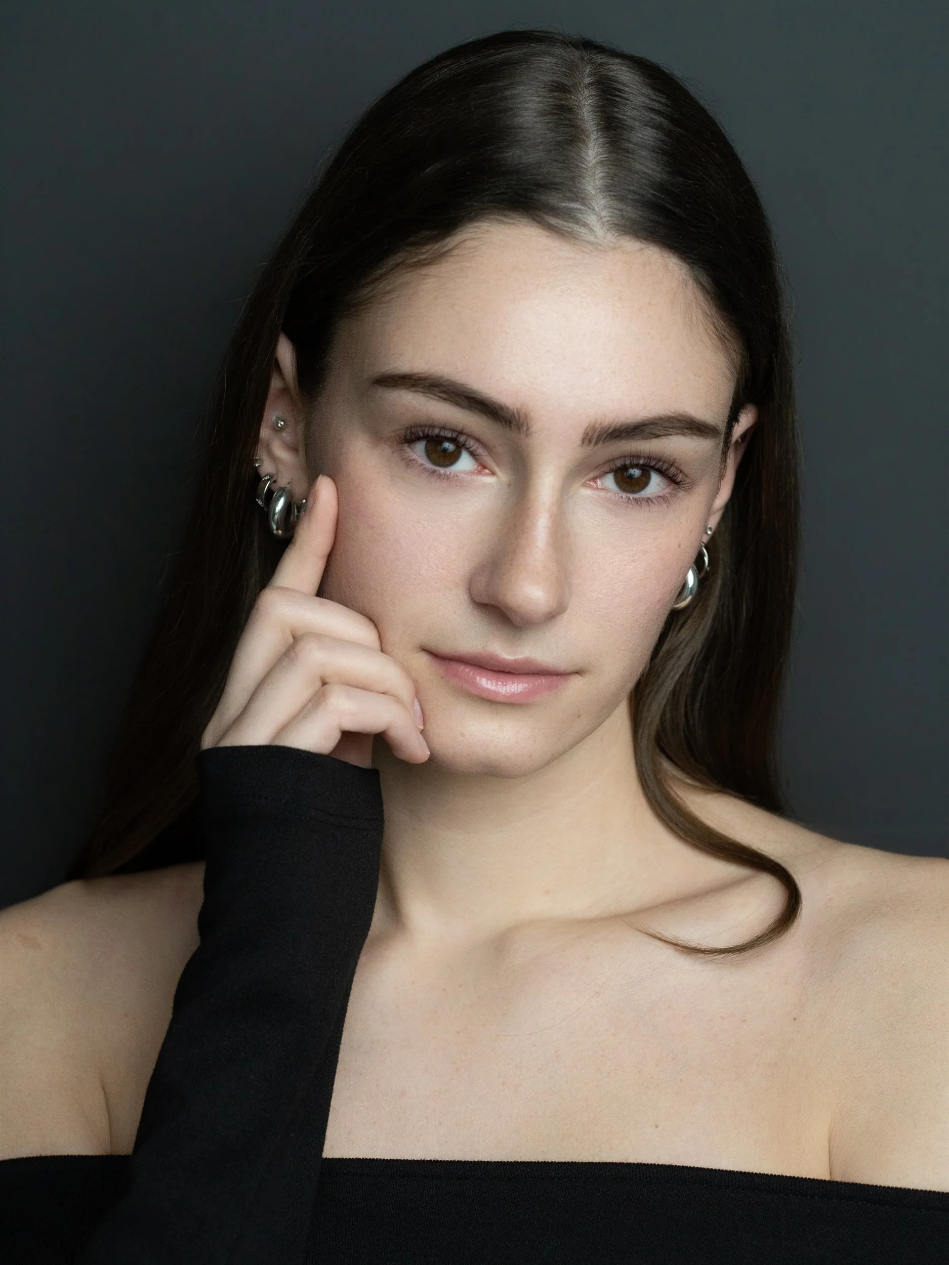 Young woman with long dark hair resting a finger against her cheek, wearing an off-the-shoulder black top and silver hoop earrings, against a dark gray background.