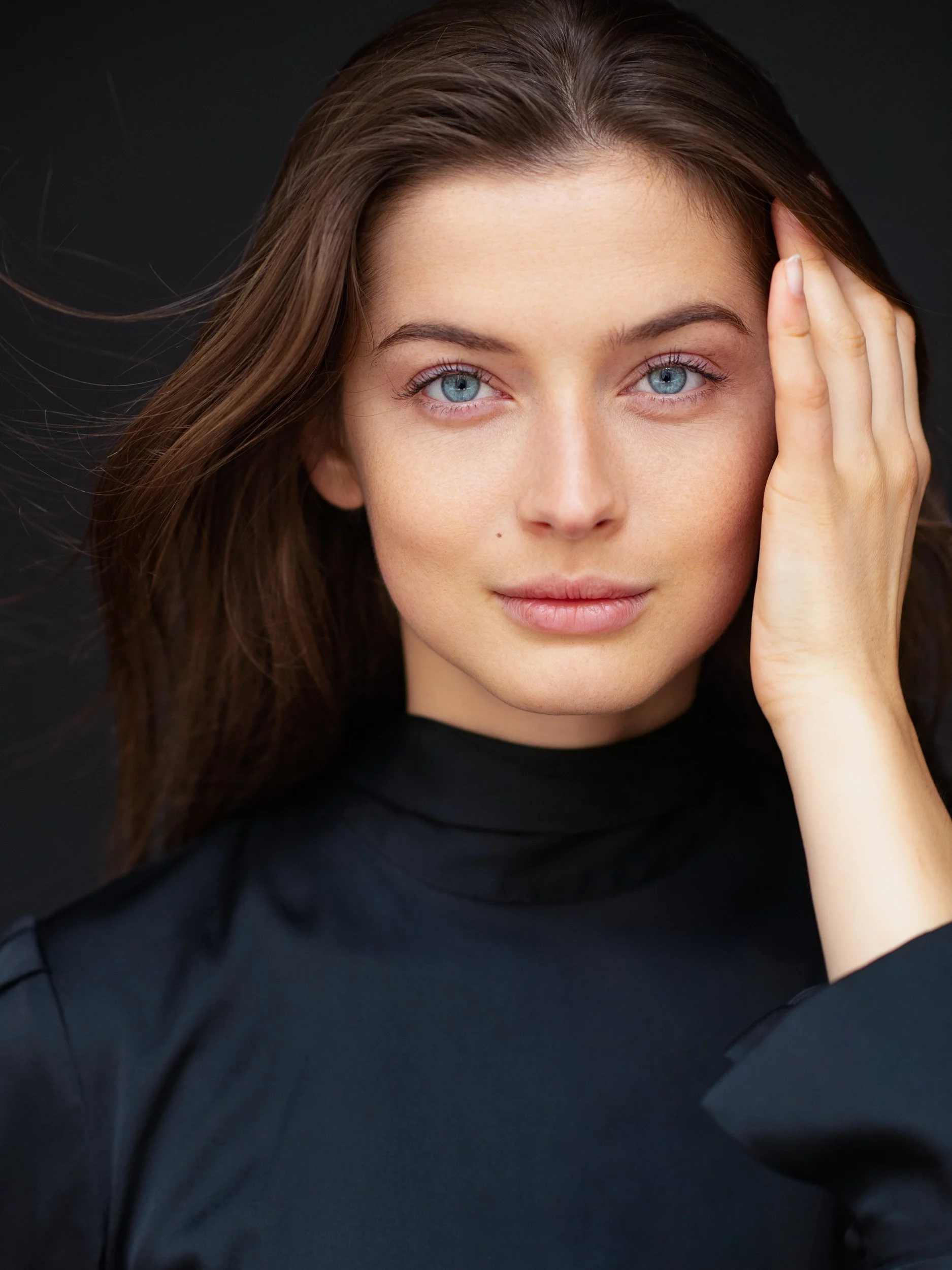 Anna with a calm, direct gaze and luminous blue eyes, hand resting at her temple, brown hair against black turtleneck and backdrop, low-key studio.