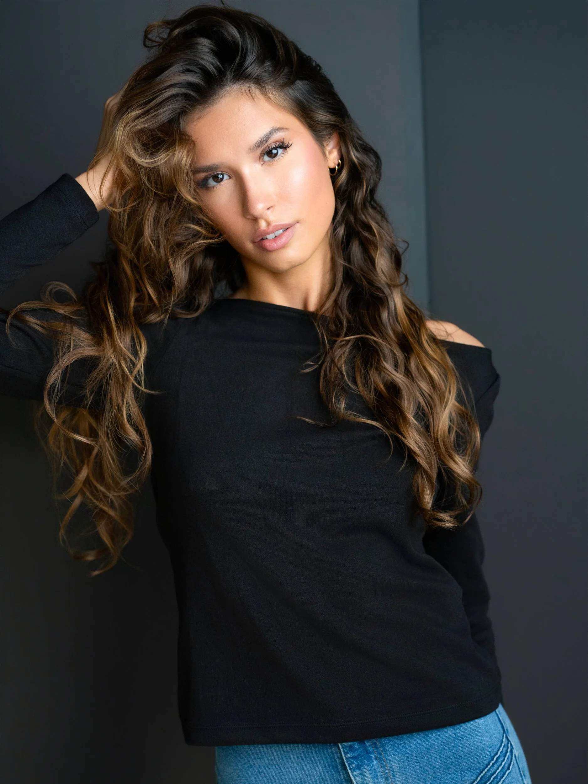Bryanna — hand in hair, lips parted, off-shoulder black top, denim, wavy balayage, low-key light on grey.