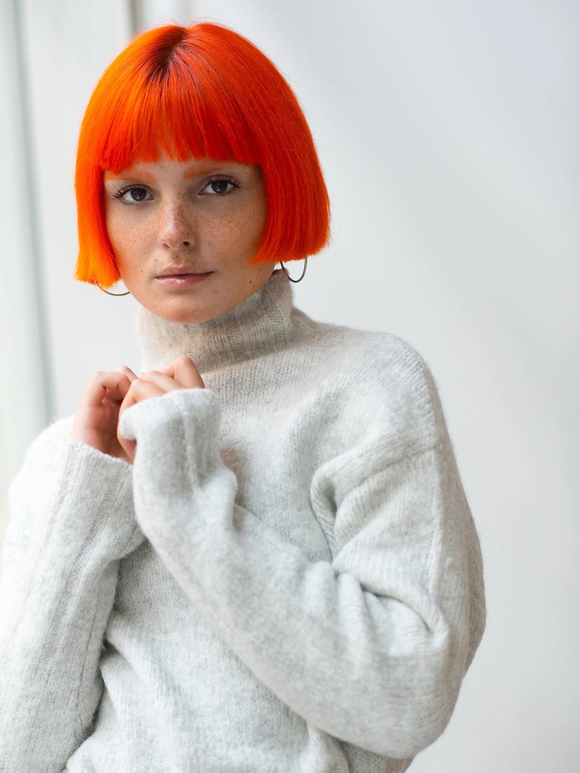 Cait gazing at the camera, vivid orange bob with blunt fringe and freckled skin, hands tucked into an oversized cream sweater, airy studio light.