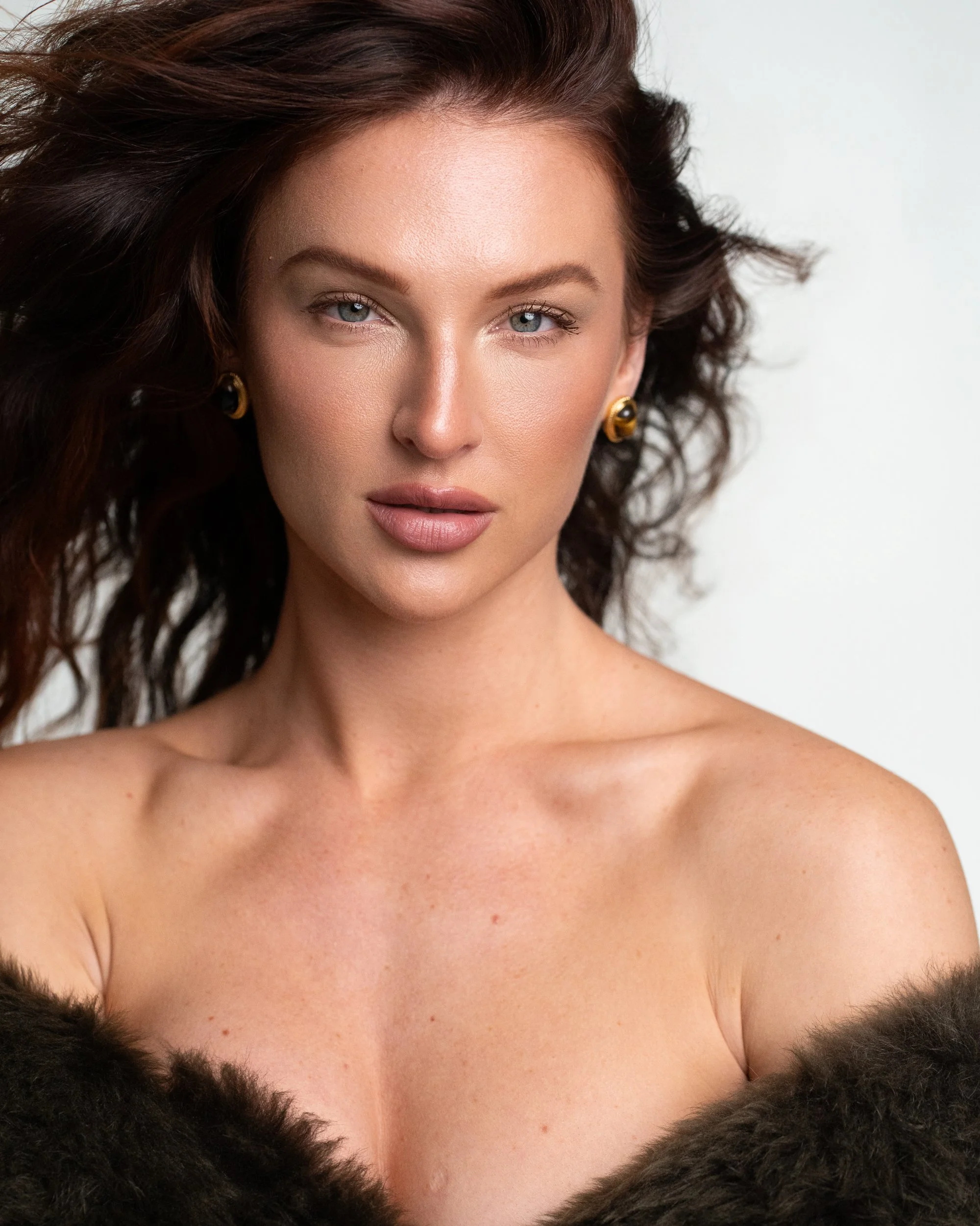 Phoebe gazing past the camera, luminous skin and tousled dark hair, gold earrings, draped fur off the shoulders, studio.