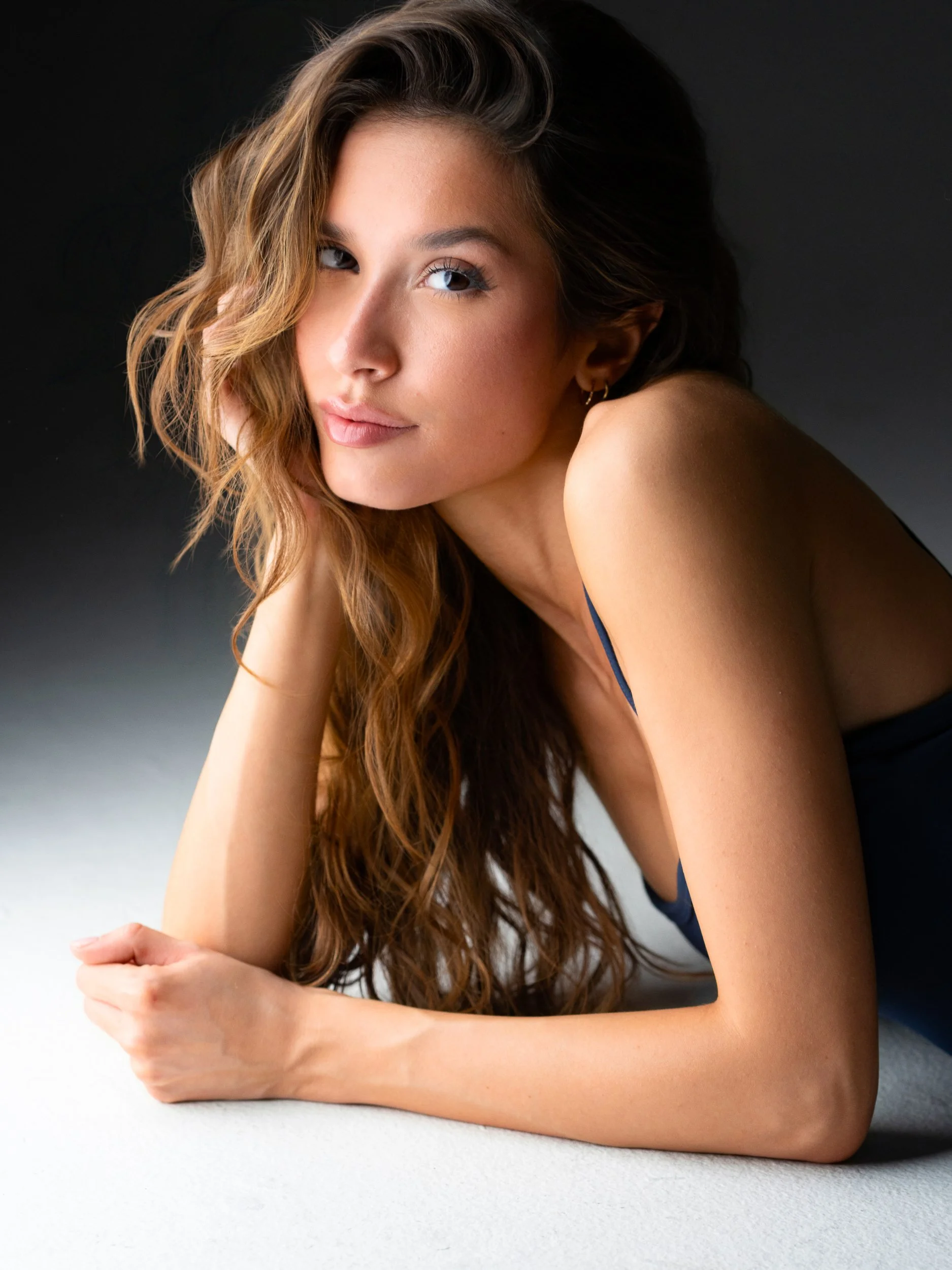 Bryanna — reclining on her forearms, direct gaze, wavy honey-brown hair, navy strap top, low-key light on dark seamless.