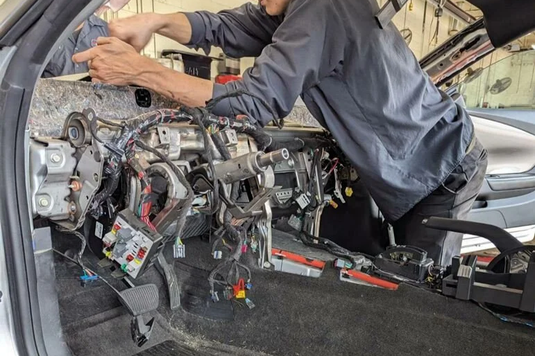 Person working on car dashboard with exposed wiring and electronic components in a garage.