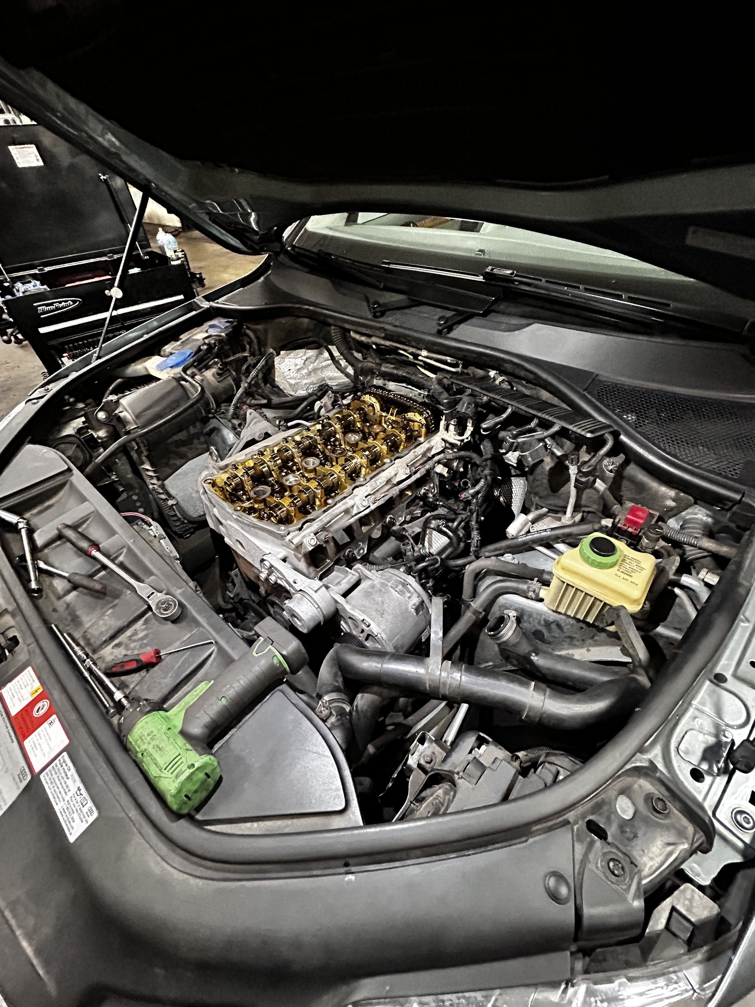 Open hood of a car showing engine with parts removed and tools nearby, indicating engine repair or maintenance in progress.