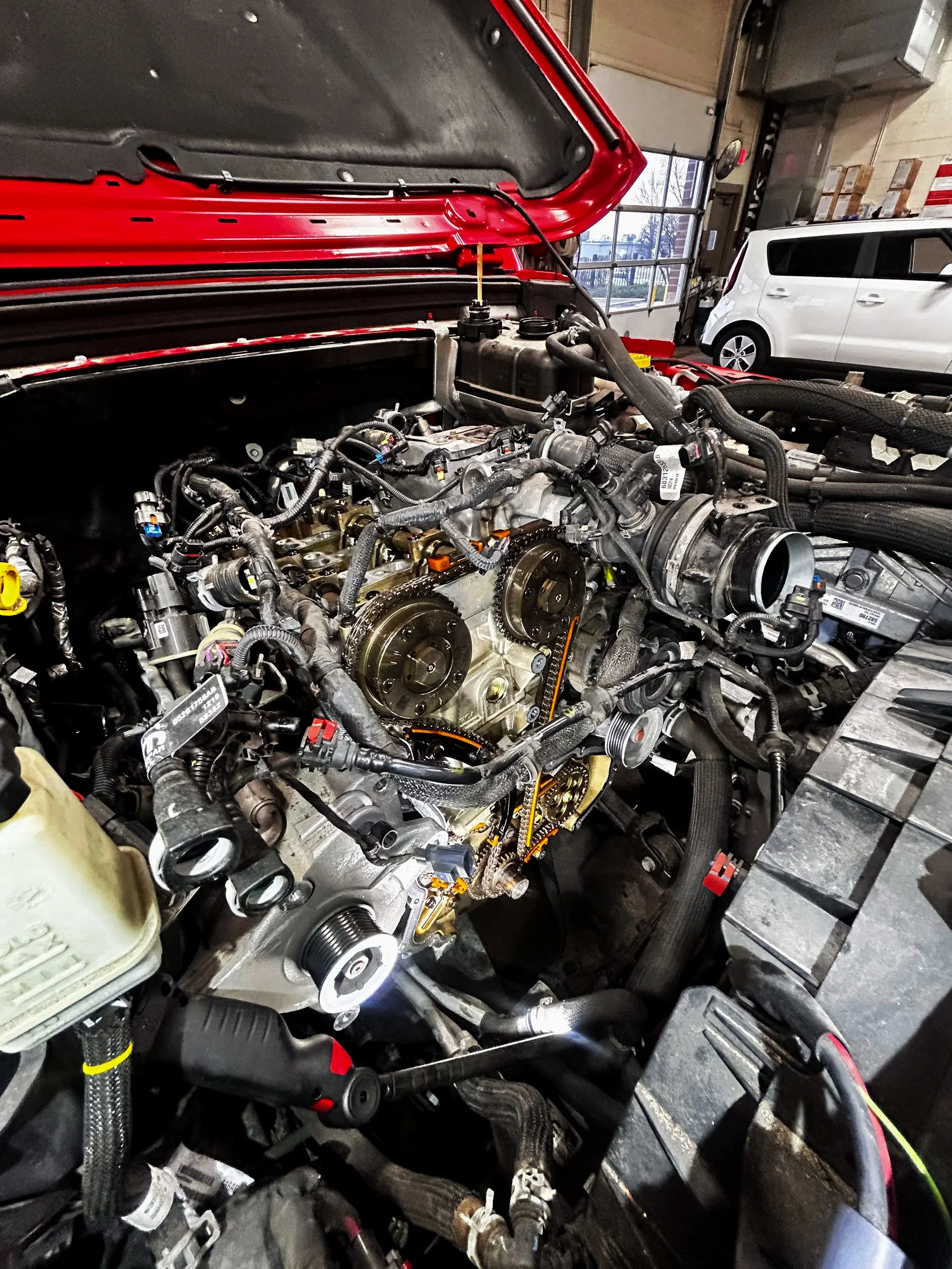Open hood of red vehicle showing engine with timing gears and various engine components in a workshop.