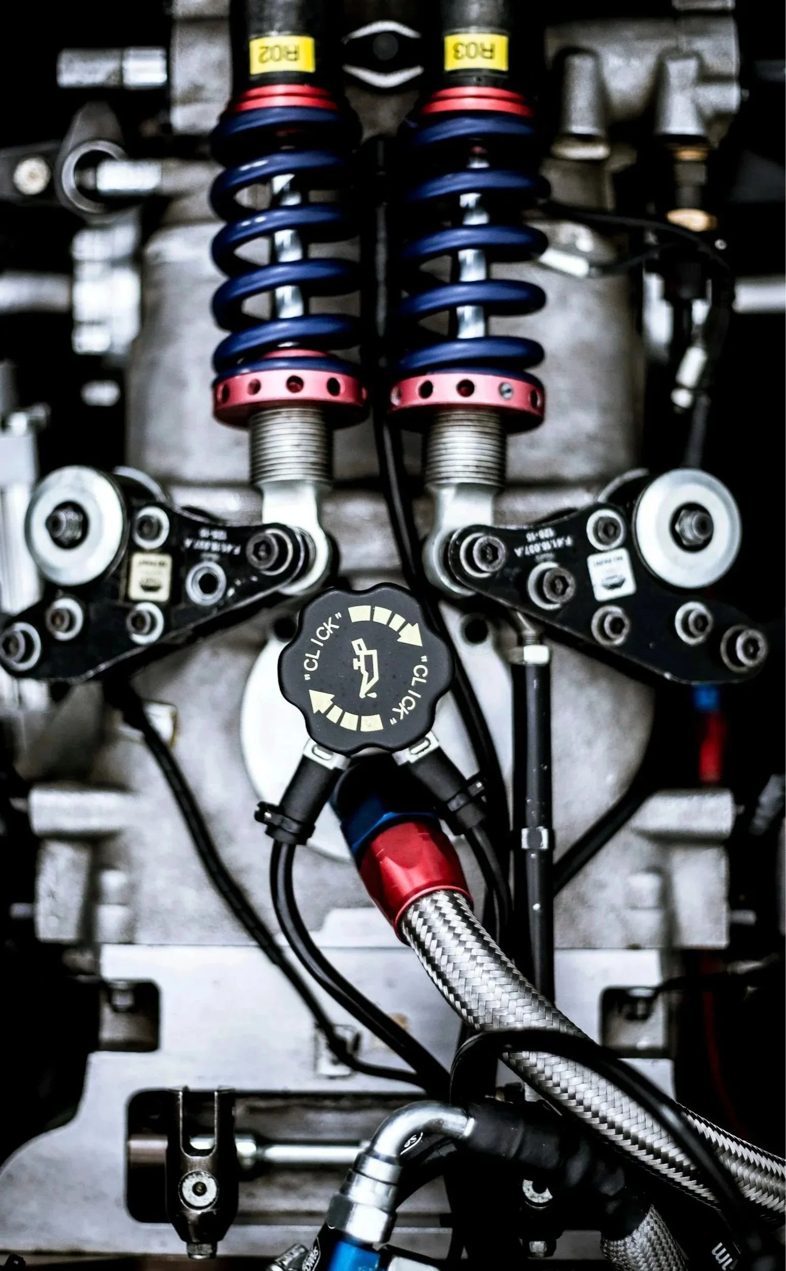 Close-up view of a racing car's suspension system, showcasing dual blue coil springs with red adjustment rings, black mounting components, and braided metal hoses connected to the engine or transmission.