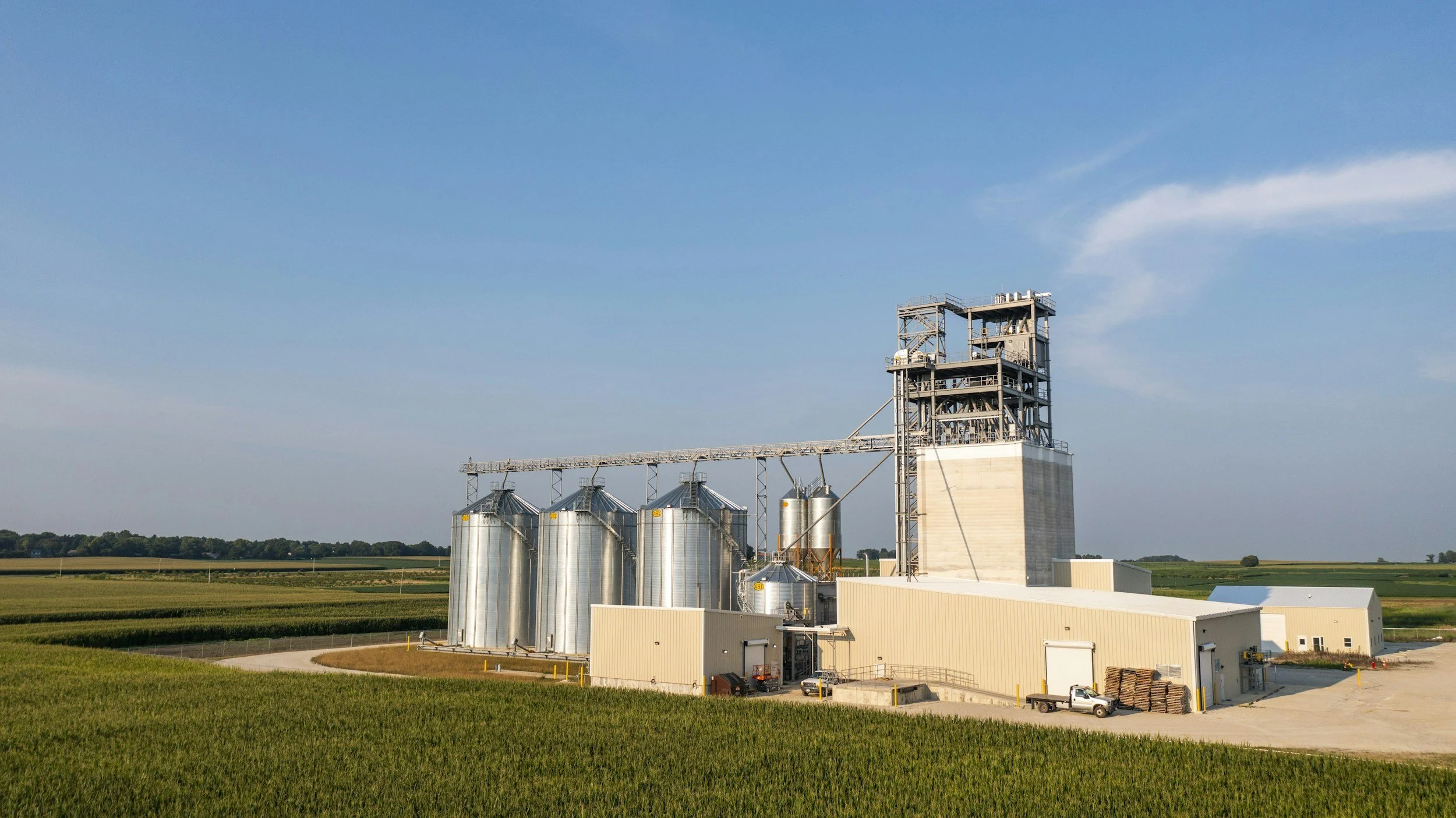 Large grain storage and processing facility in a rural area with fields and clear blue sky.
