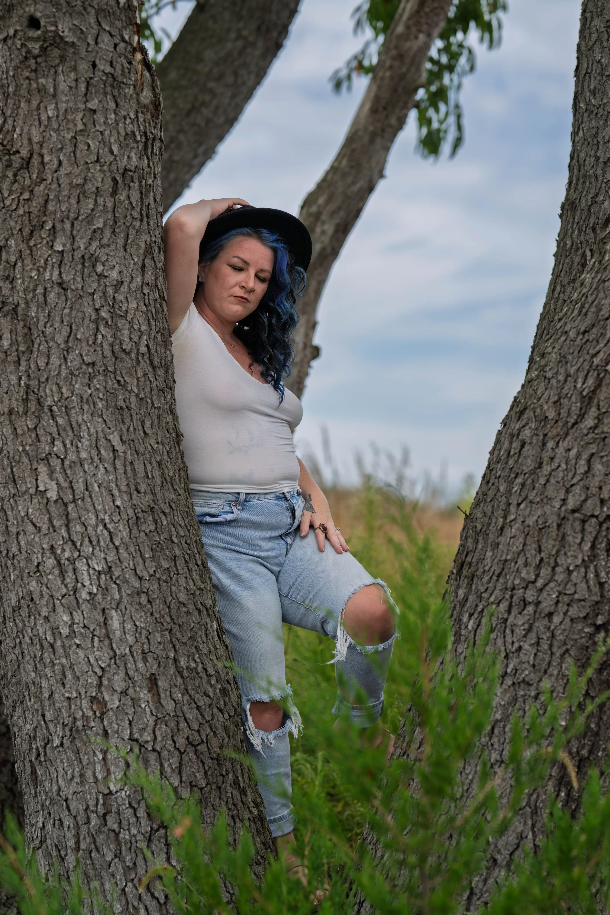 A woman with black and blue hair wearing a white t-shirt, ripped jeans, and a black hat leans against the trunk of a tree outdoors during the daytime.