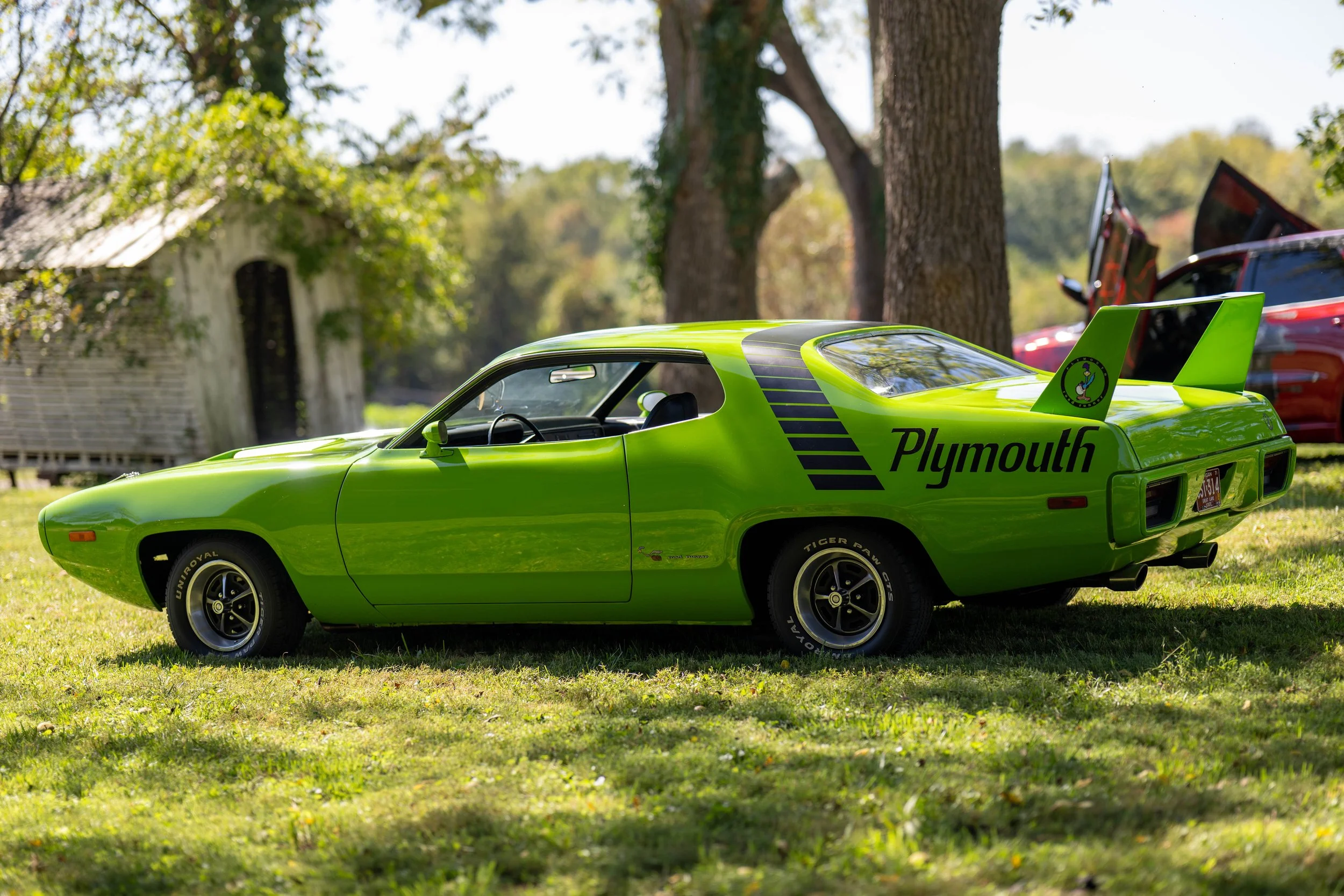 A bright green vintage Plymouth race car with black stripes and a large rear wing on a grassy area with trees and other cars in the background.