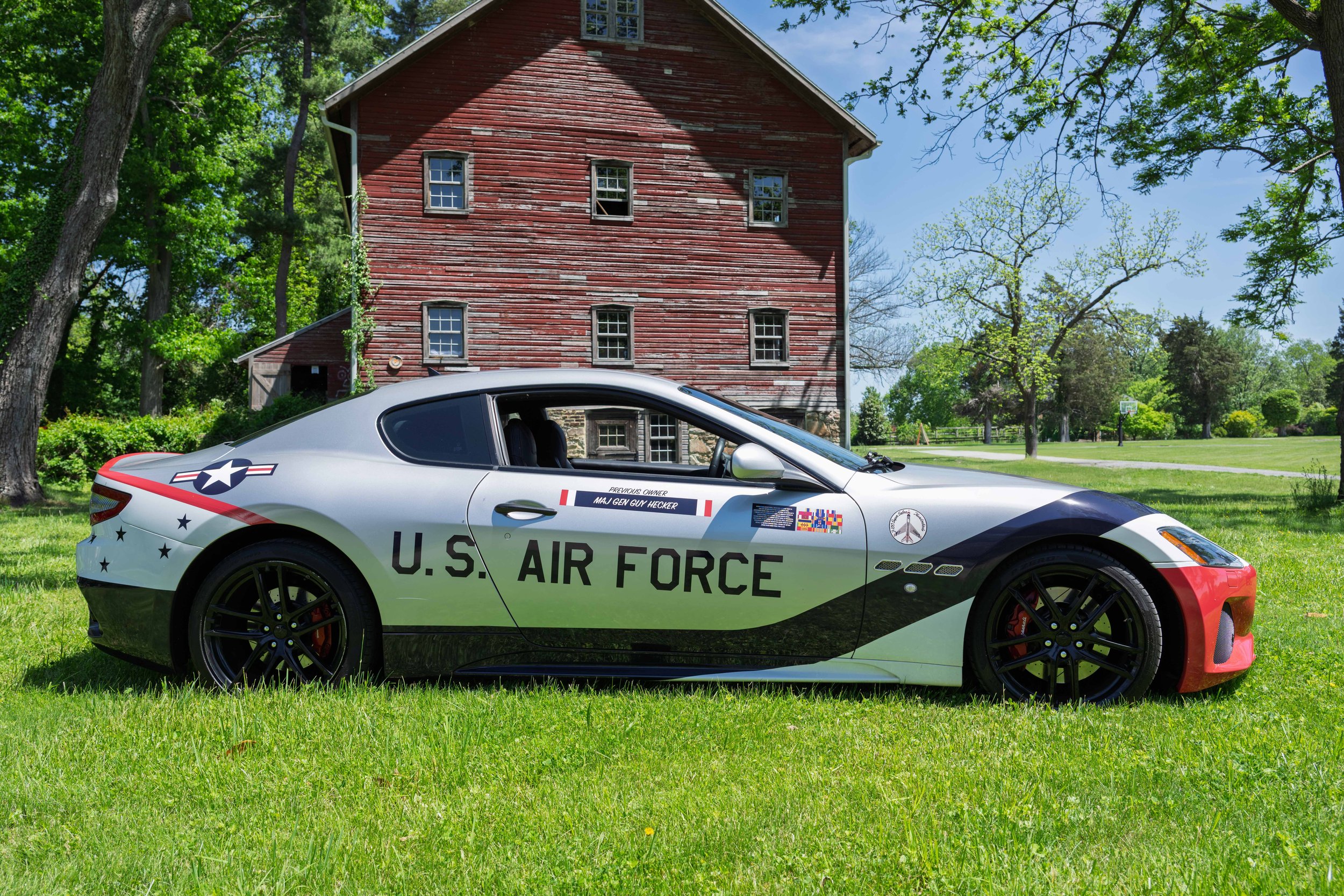 U.S. Air Force-themed sports car parked on a grassy area in front of a red wooden farm building with trees and a playground in the background.