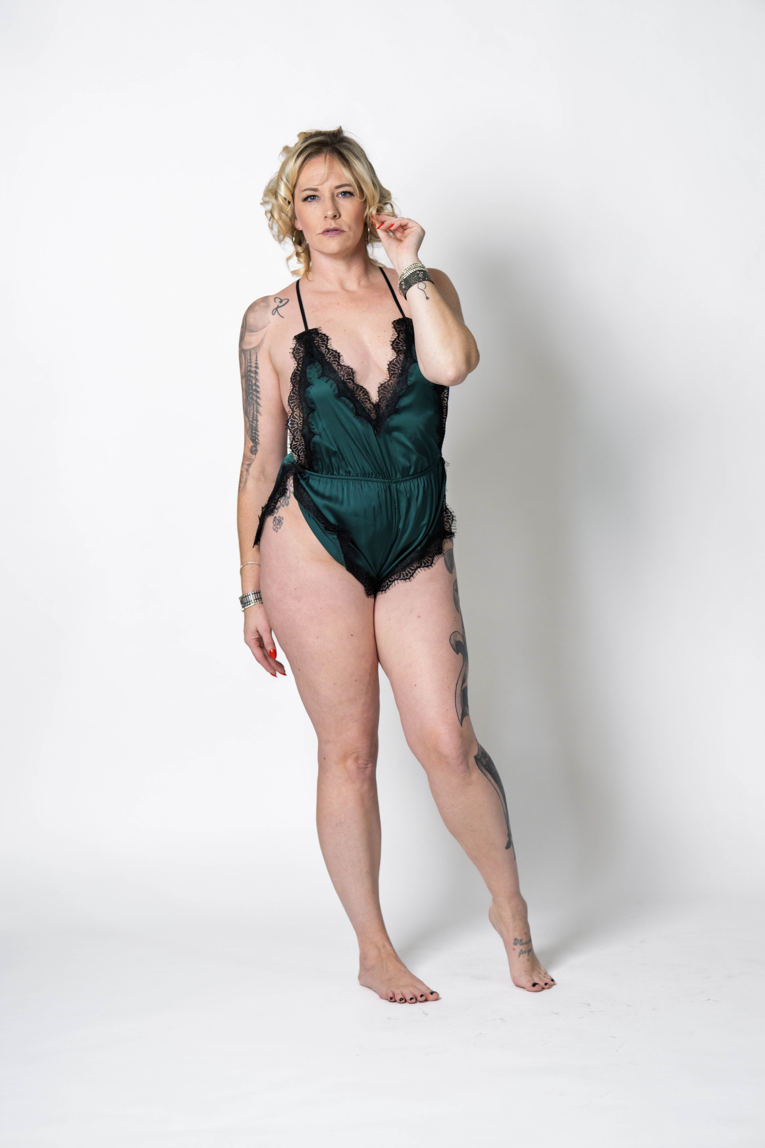 A woman with blonde hair, tattoos, wearing a dark green satin lingerie piece with black lace trim, standing barefoot against a plain white background.