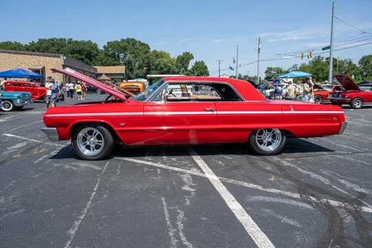 A red vintage car with its hood open parked in a parking lot during a car show.