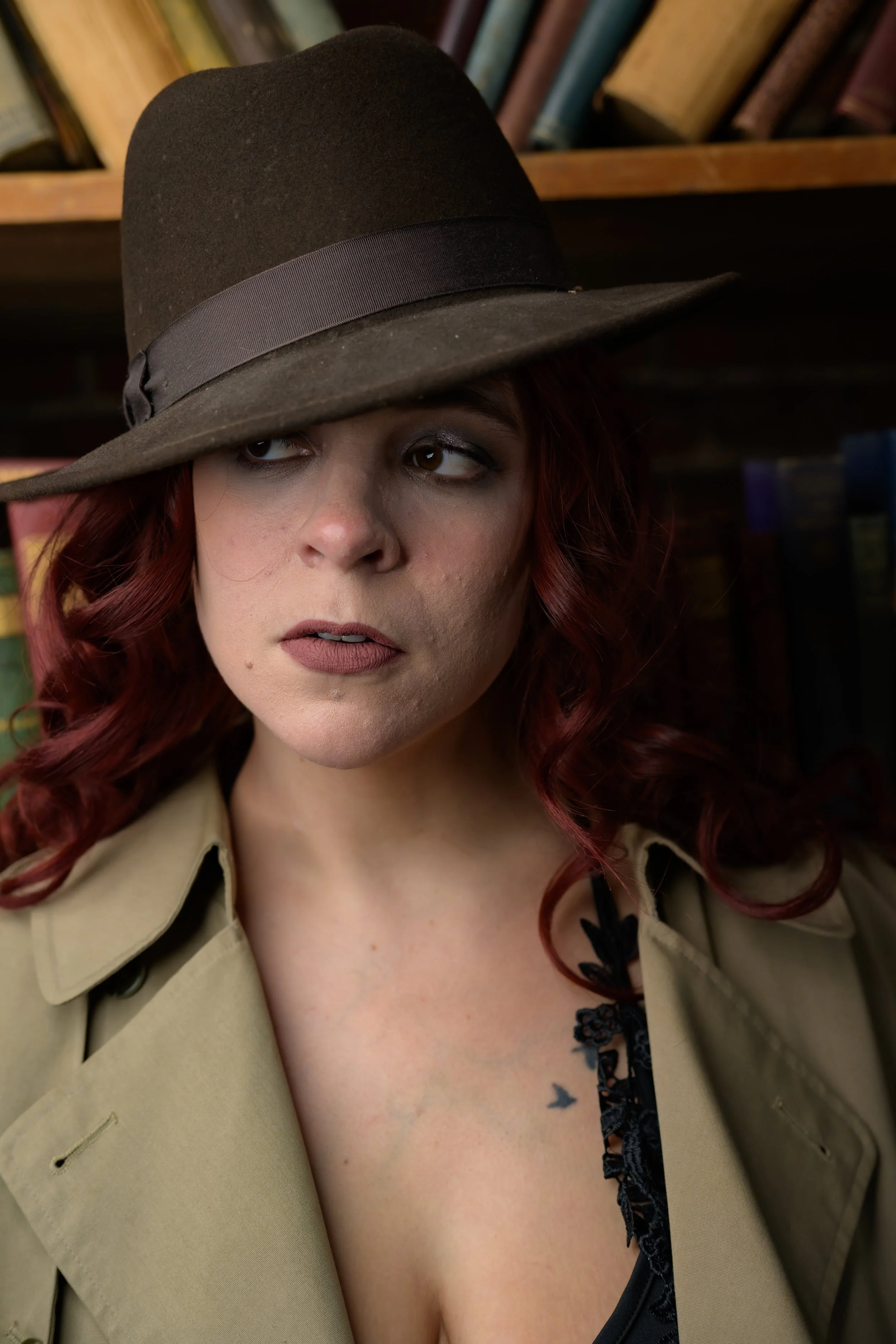 A woman with red hair wearing a tan trench coat and a large brown felt hat with a ribbon, standing in front of a bookshelf.