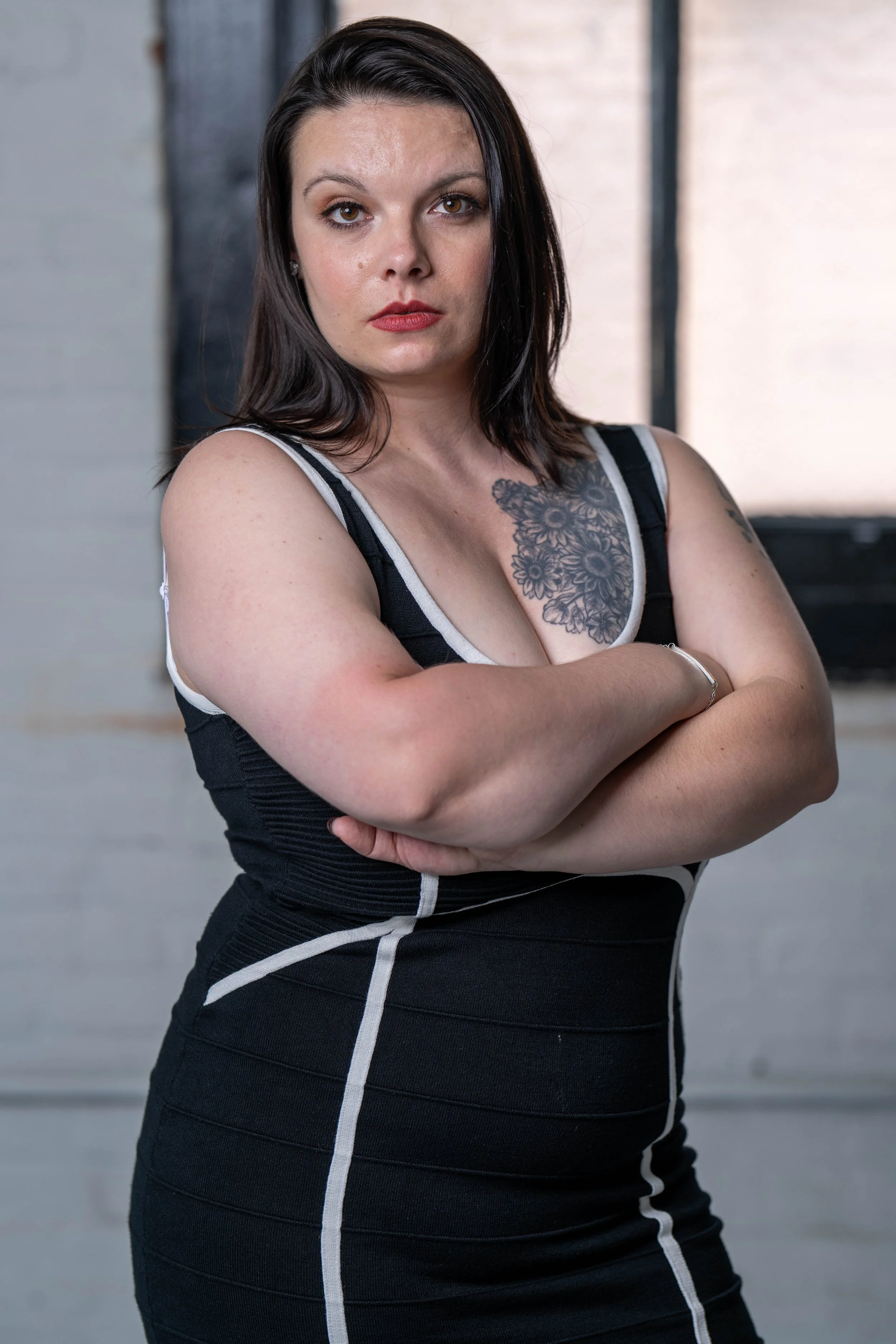 A woman with dark brown hair and light skin, wearing a black dress with white trim, standing with her arms crossed in front of her chest in front of a blurred background with a window.