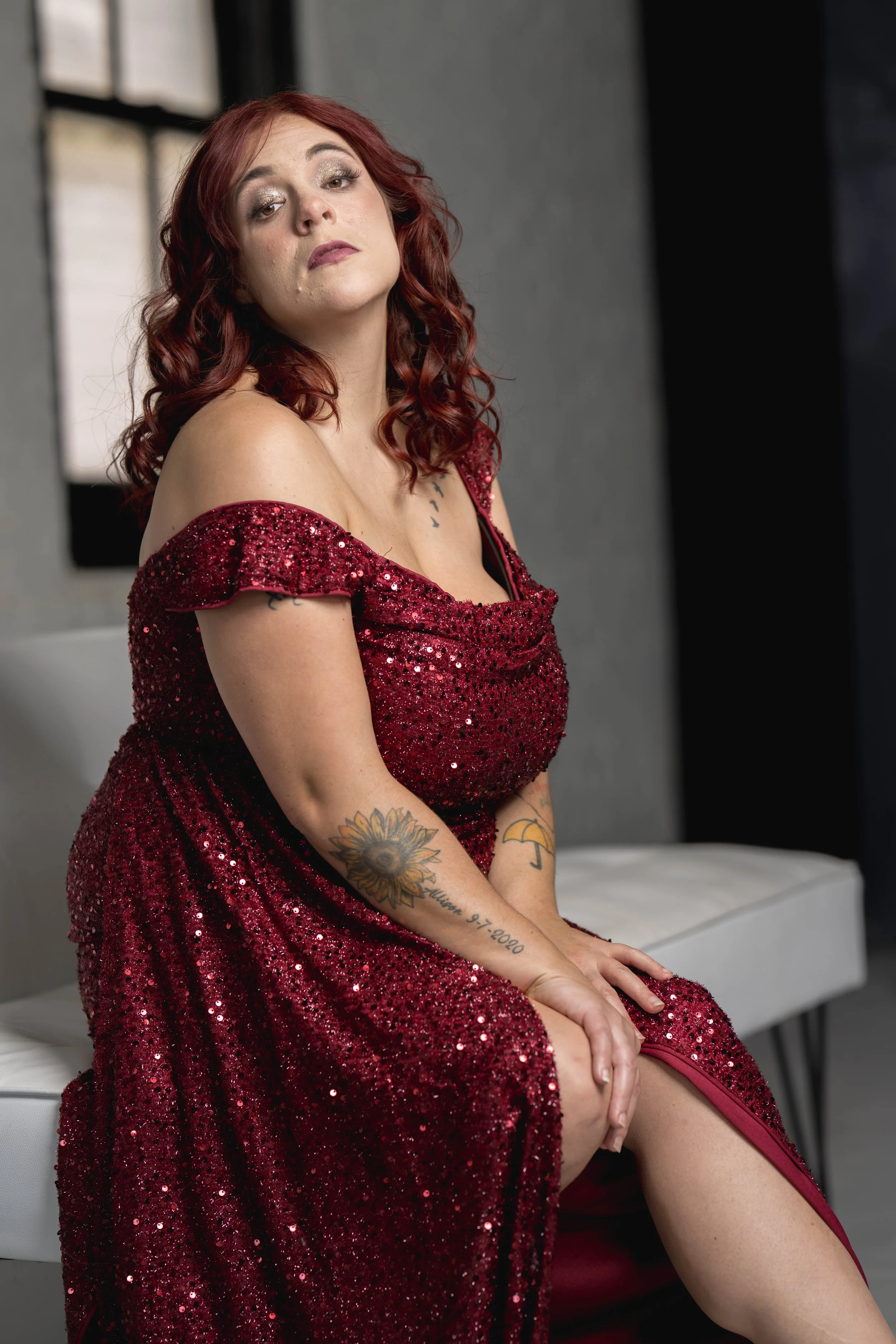A woman with red curly hair sitting on a bench in a red sequined off-shoulder dress, with tattoos on her arms and chest, in a modern room with dark walls and a window.