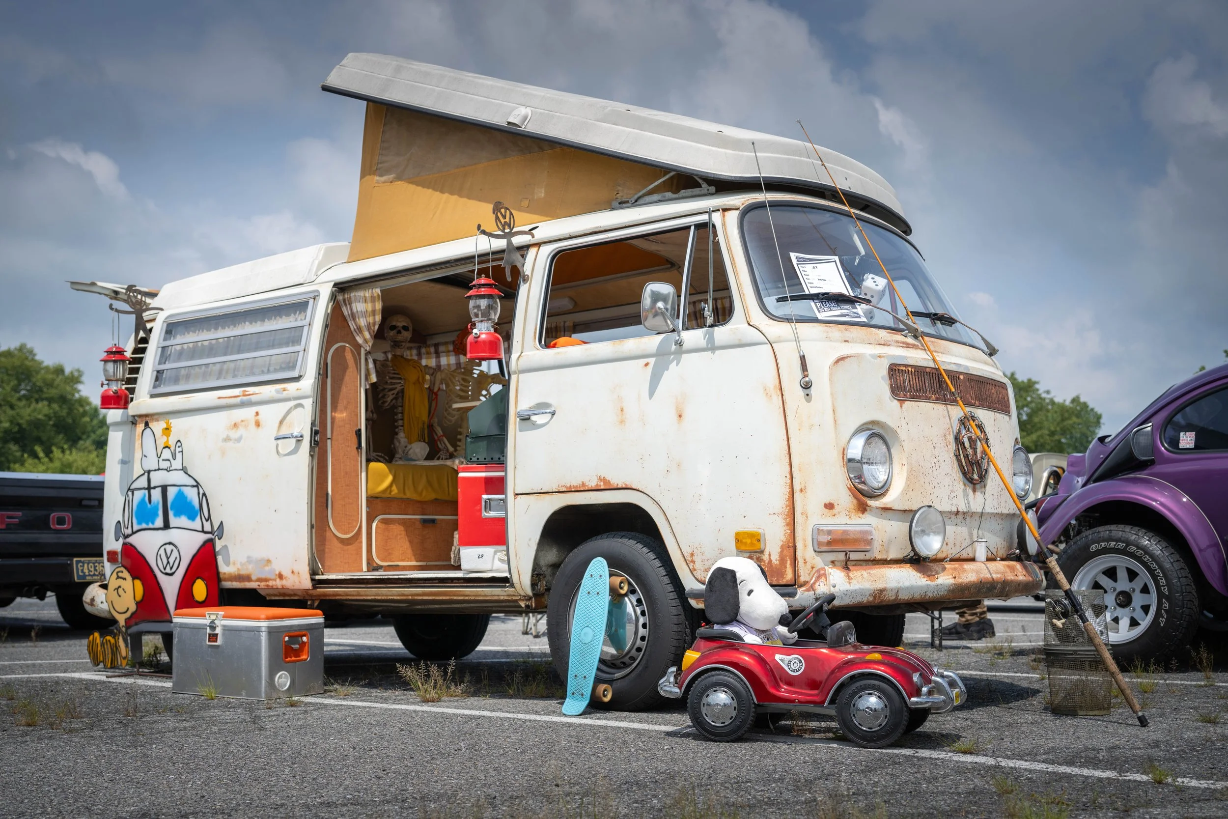 A vintage Volkswagen bus with rust spots and a partially retracted pop-up roof, parked in a lot with other vehicles. The bus has a cartoonish decal of a camper van with sunglasses on the side. Items like a skateboard, a small red toy car with a dog p