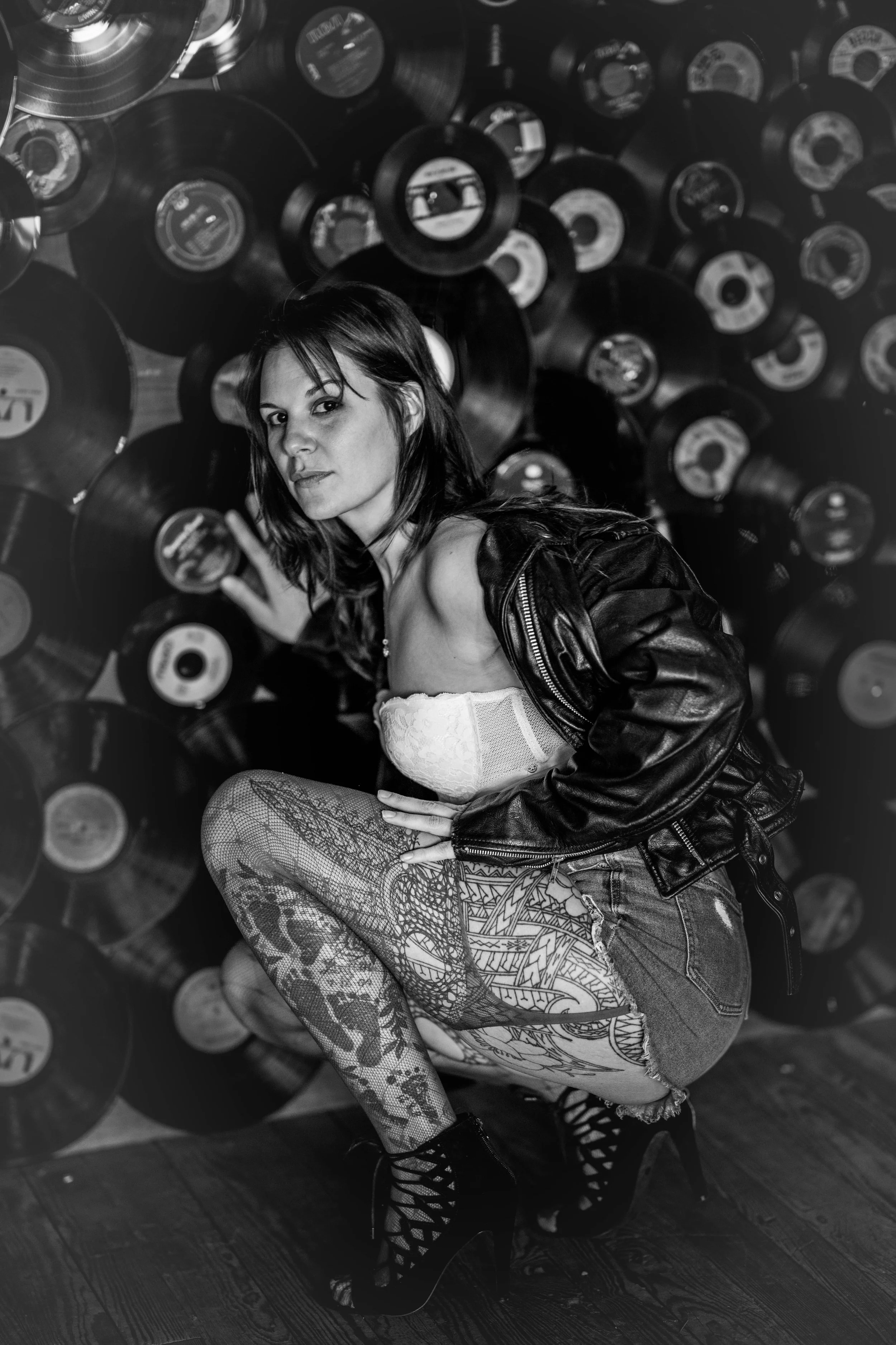 A woman with tattoos, wearing a leather jacket, lace top, ripped jeans, and high heels, crouching in front of a wall of vinyl records.