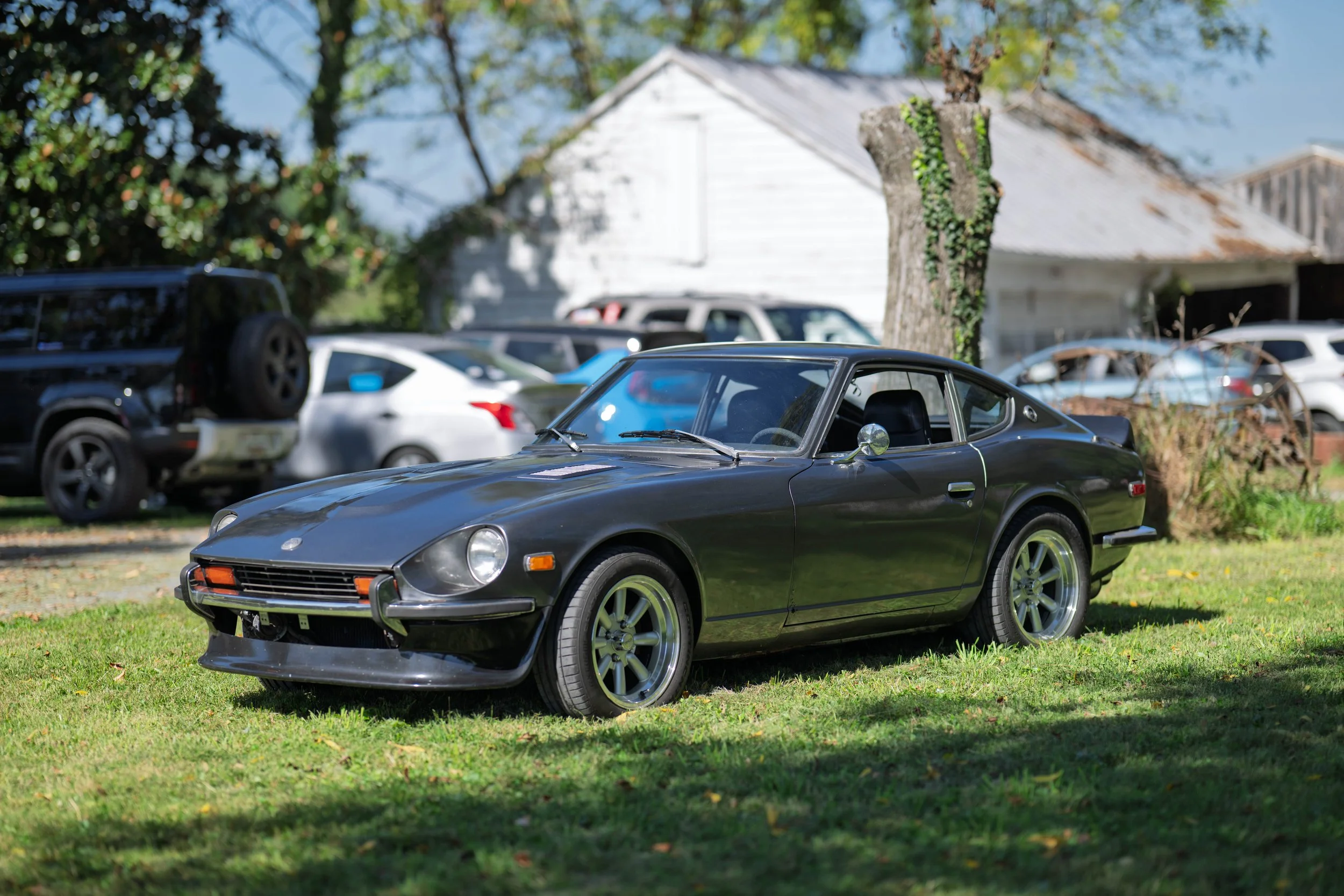 A black vintage sports car parked on a grassy area with trees and a white house in the background.