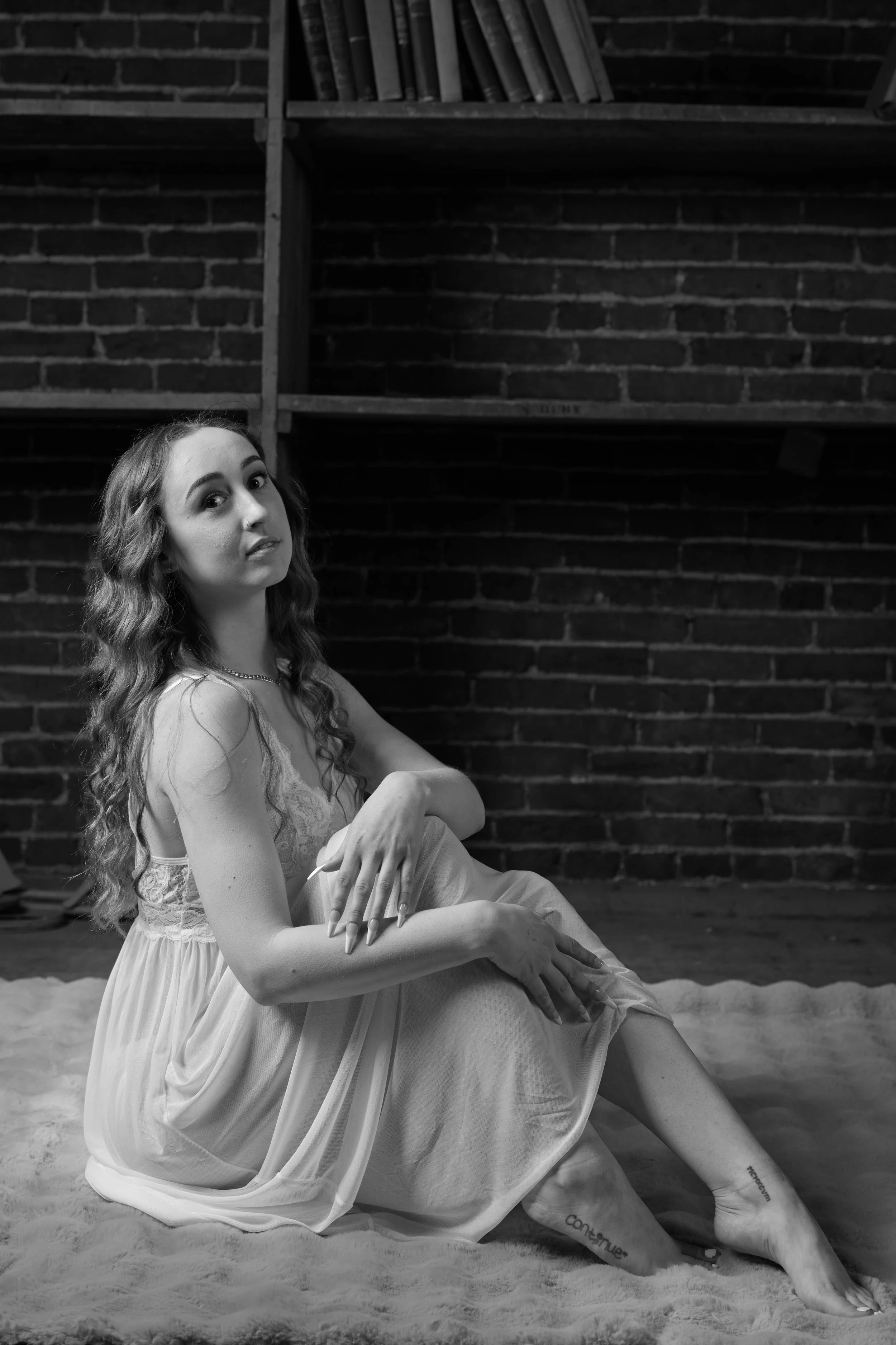 A black and white photo of a young woman with long, wavy hair sitting on a fluffy rug in front of a brick wall and bookshelves. She is wearing a light, sleeveless dress and necklace, with her arms crossed over her knees and looking towards the camera