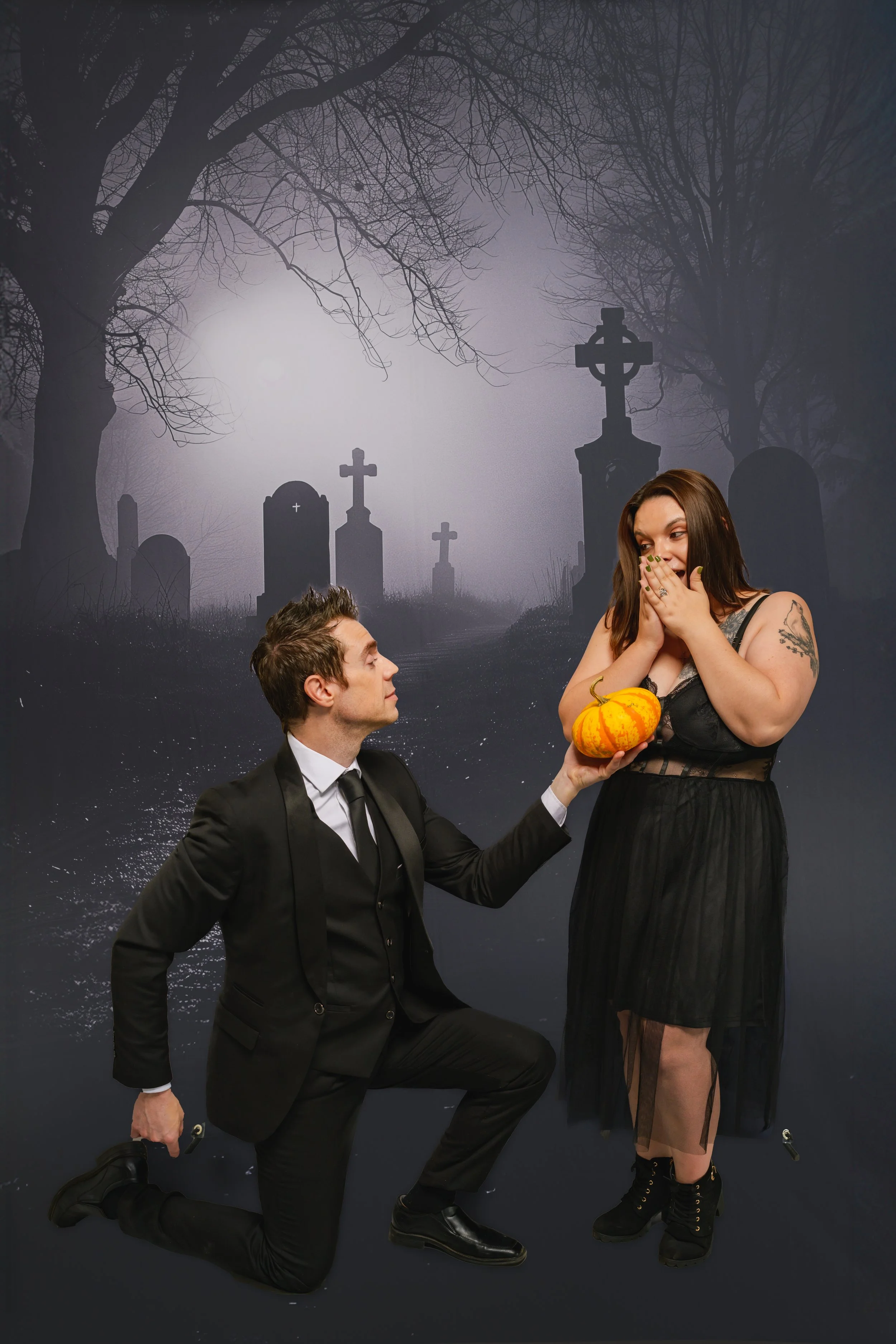 A man in a black suit on one knee proposing to a woman dressed in black with a pumpkin in her hand, set against a dark, spooky cemetery background with tombstones and leafless trees.