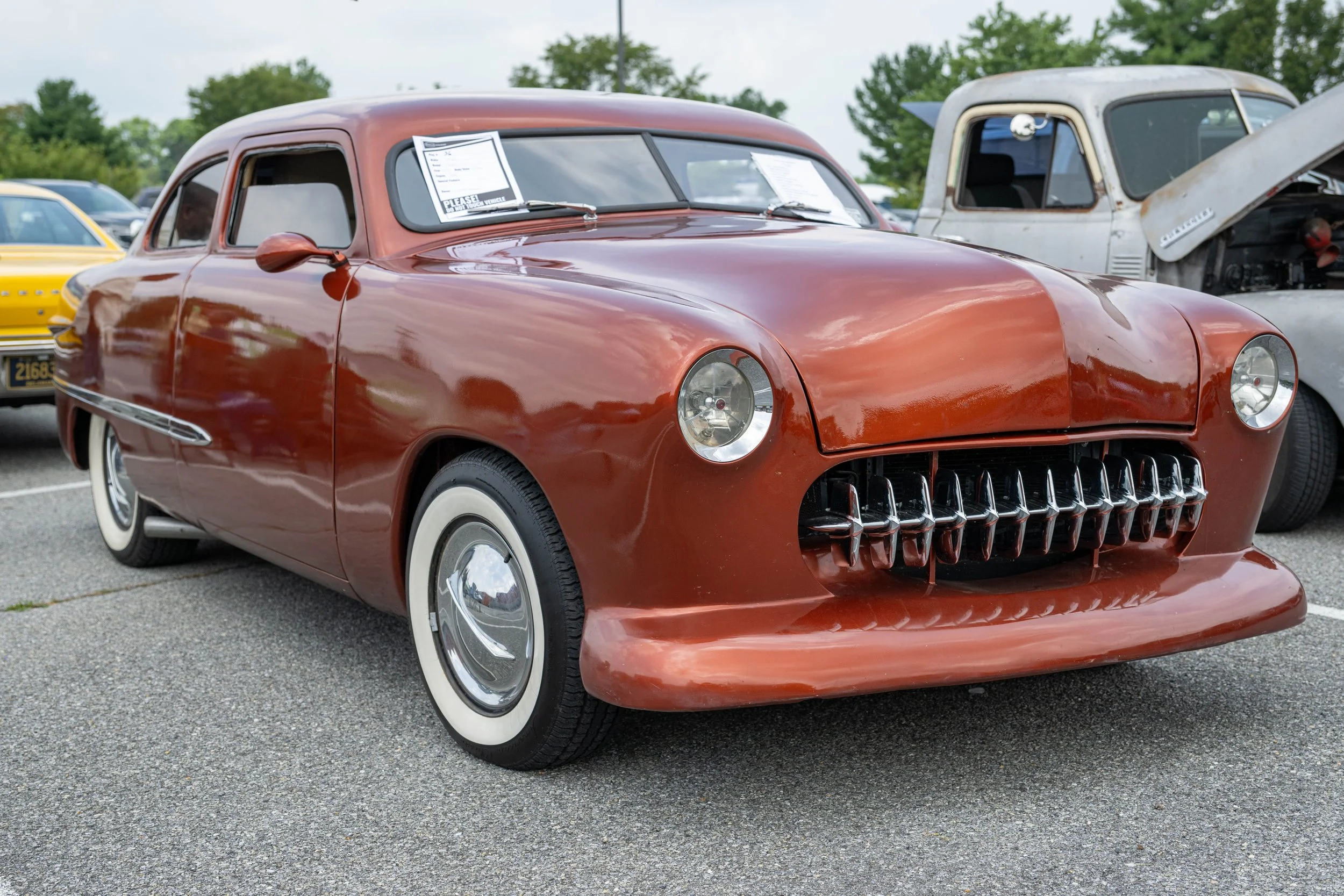 Red vintage car with unique front grille at a car show, surrounded by other classic vehicles, with trees and overcast sky in background.