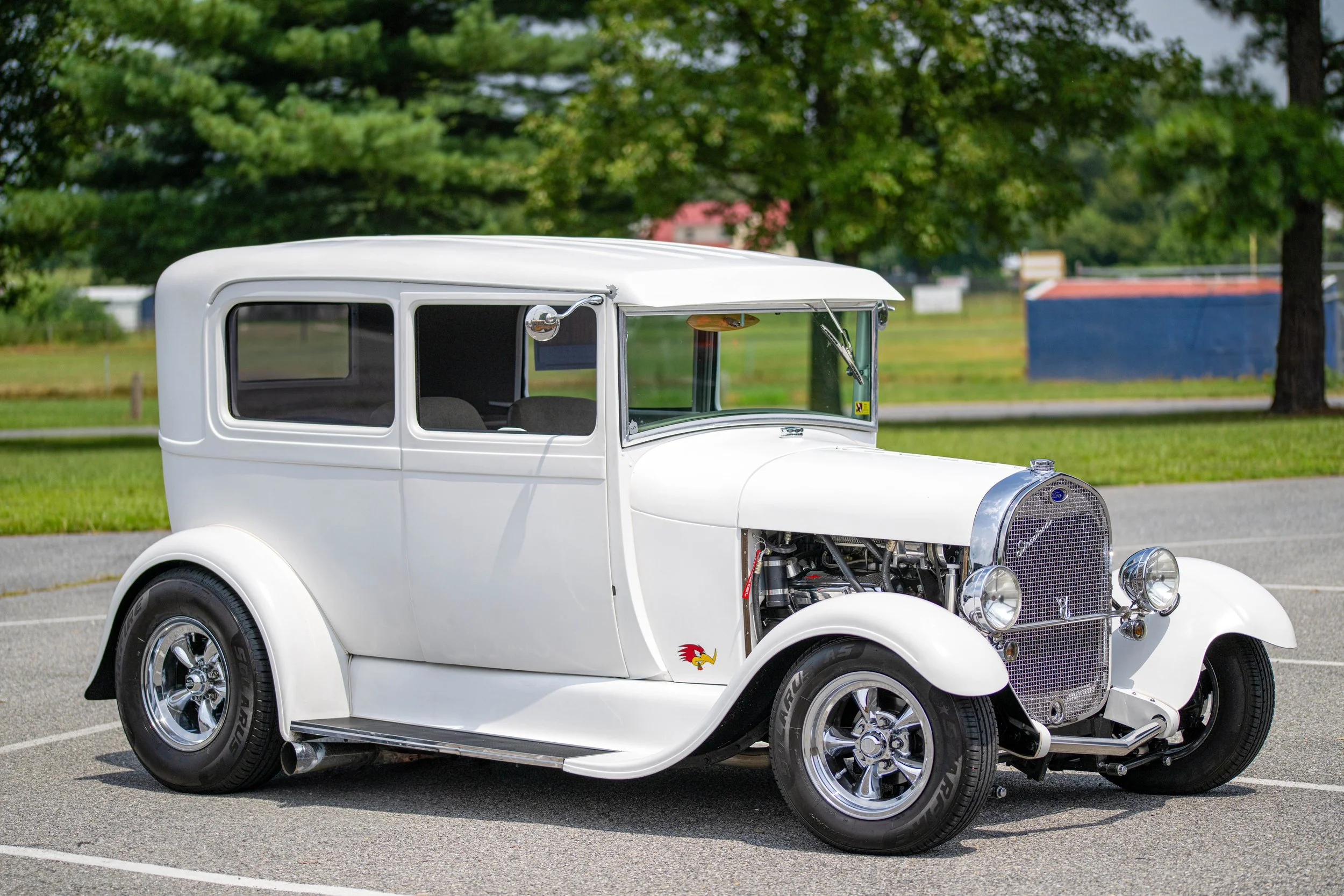 A vintage white classic car parked on a paved lot with green trees in the background.