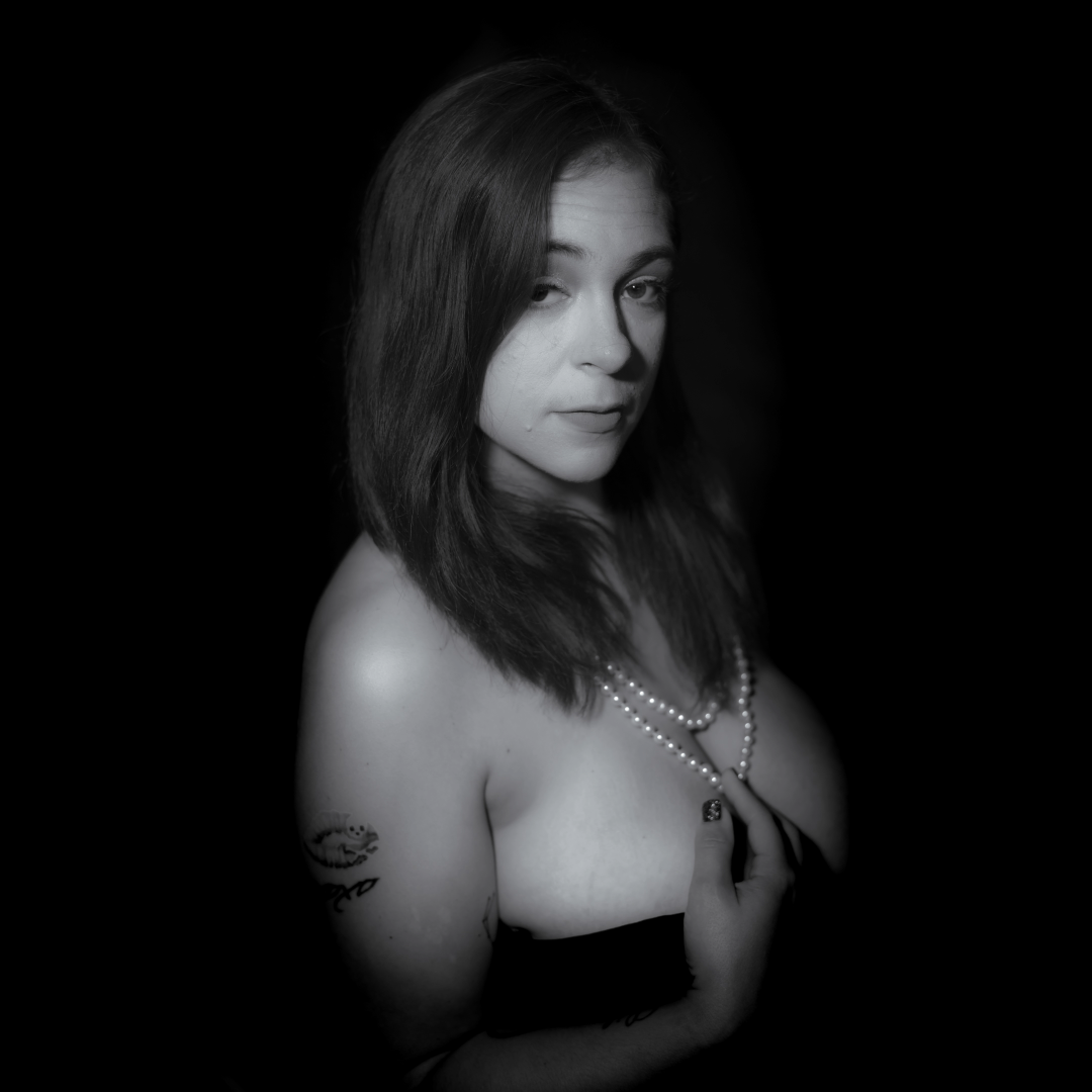 Black and white portrait of a woman with shoulder-length hair, gazing at the camera with one eye slightly closed, holding her hands over her chest, wearing pearl necklaces and a strapless top, with tattoos visible on her left arm.