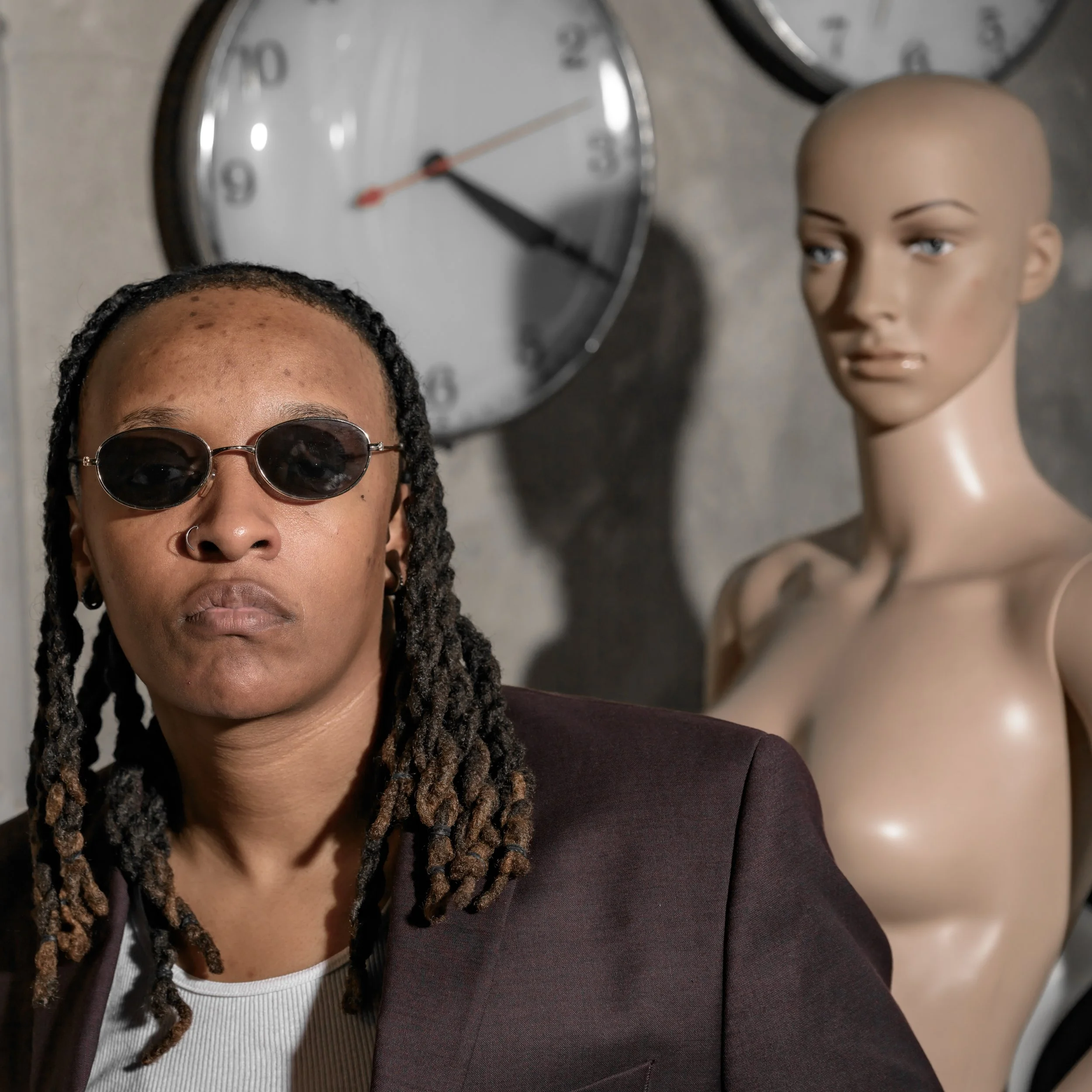 A person with dreadlocks wearing sunglasses and a dark blazer, standing in front of a wall clock and a female mannequin with a bald head.