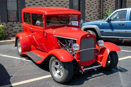 Red vintage car parked in a lot, with an exposed engine, modern silver wheels, and a boxy shape, seen beside a blue vehicle.