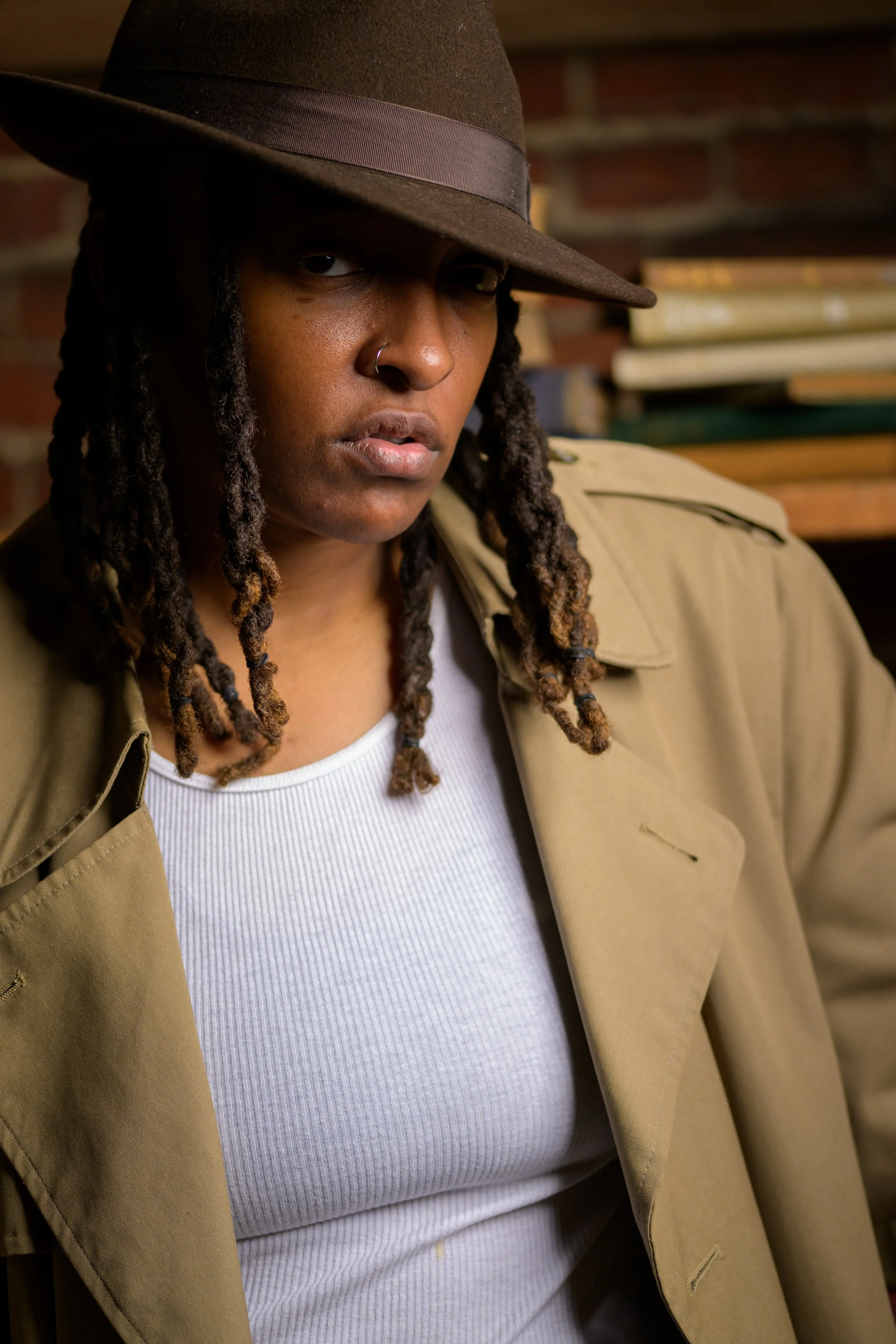 A woman with long dreadlocks, wearing a dark brown fedora hat, a beige trench coat, and a white ribbed shirt, standing against a brick wall background.