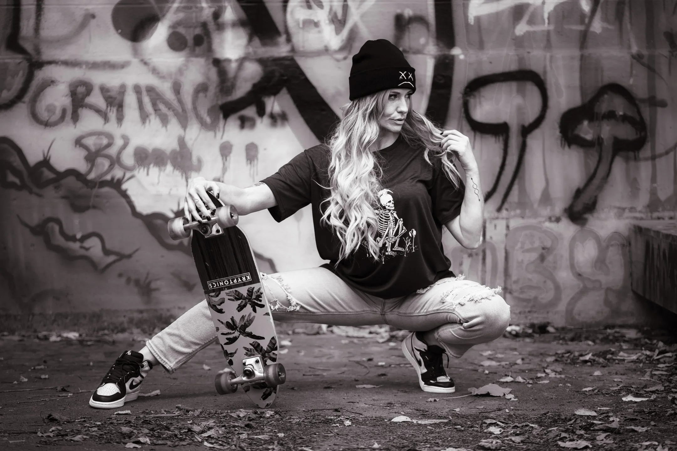 A woman with long wavy hair, wearing a beanie and skateboarder clothes, crouches on the ground holding a skateboard, with a graffiti-covered wall in the background.