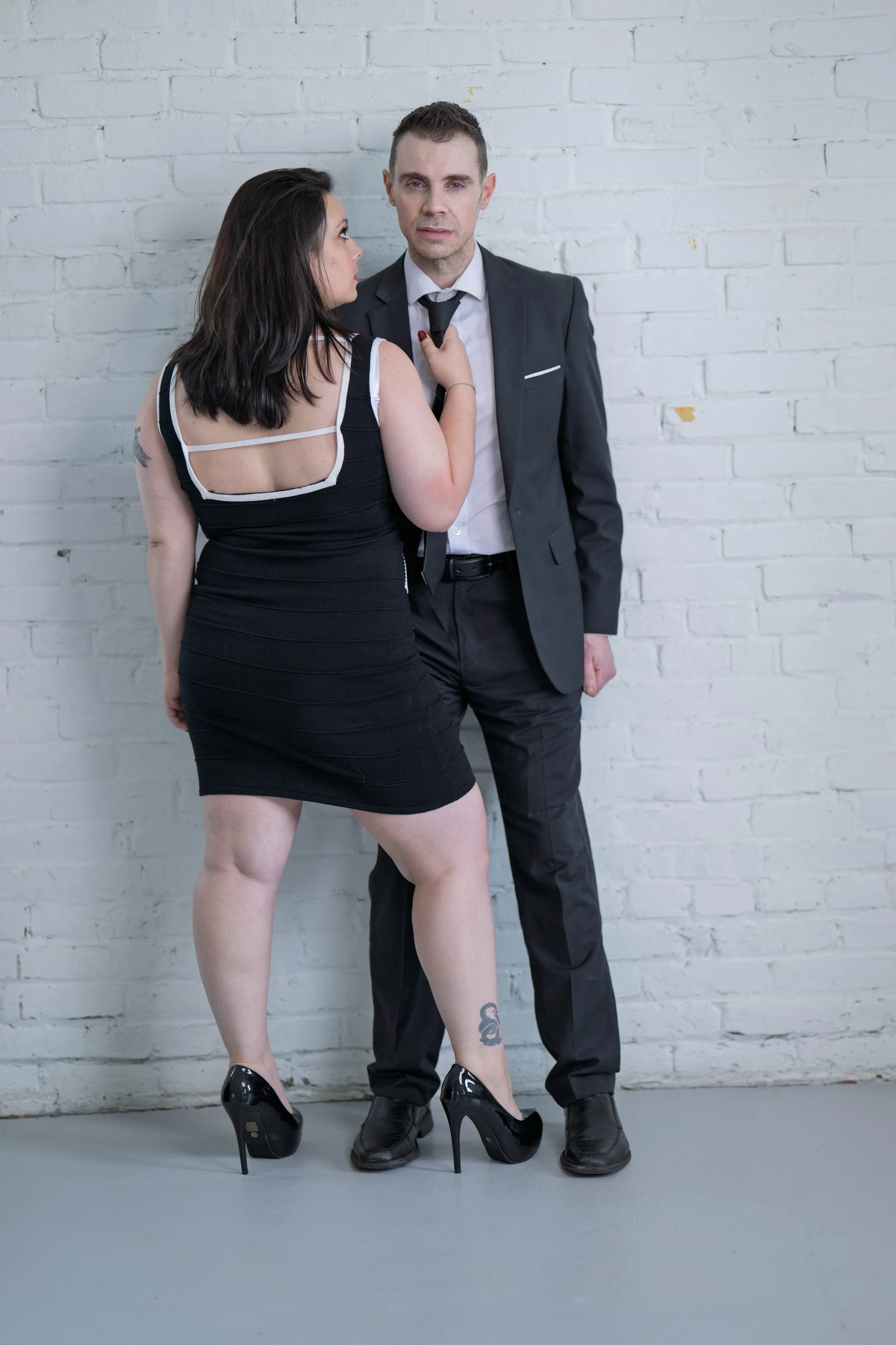 A woman in a black dress and high heels is standing close to a man in a black suit and tie, against a white brick wall. The woman is looking at the man, and the man is looking at the camera.