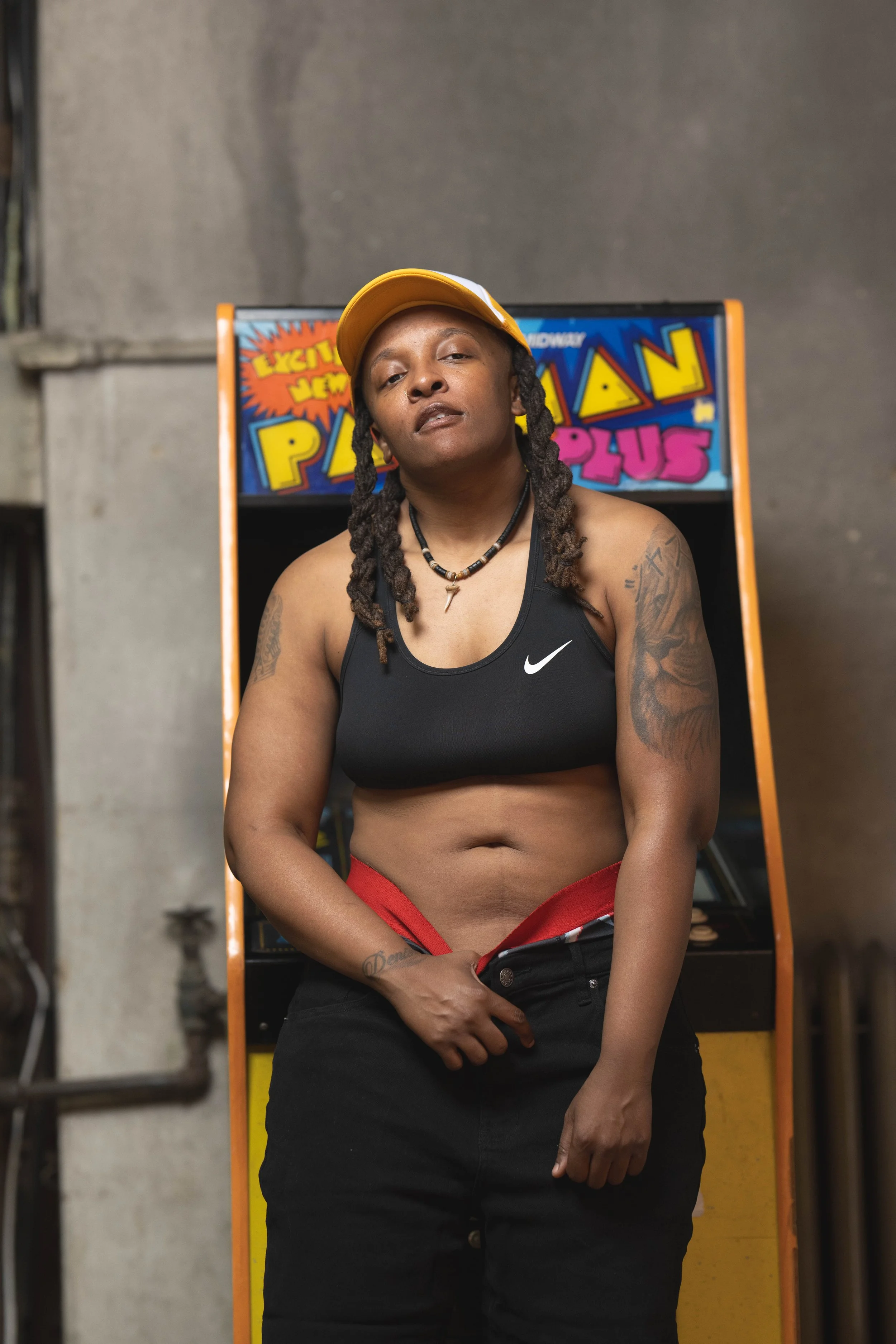 A woman with earrings, tattoos, and a necklace standing in front of an arcade game machine, wearing a black sports bra and black pants, with a yellow cap tilted back on her head.