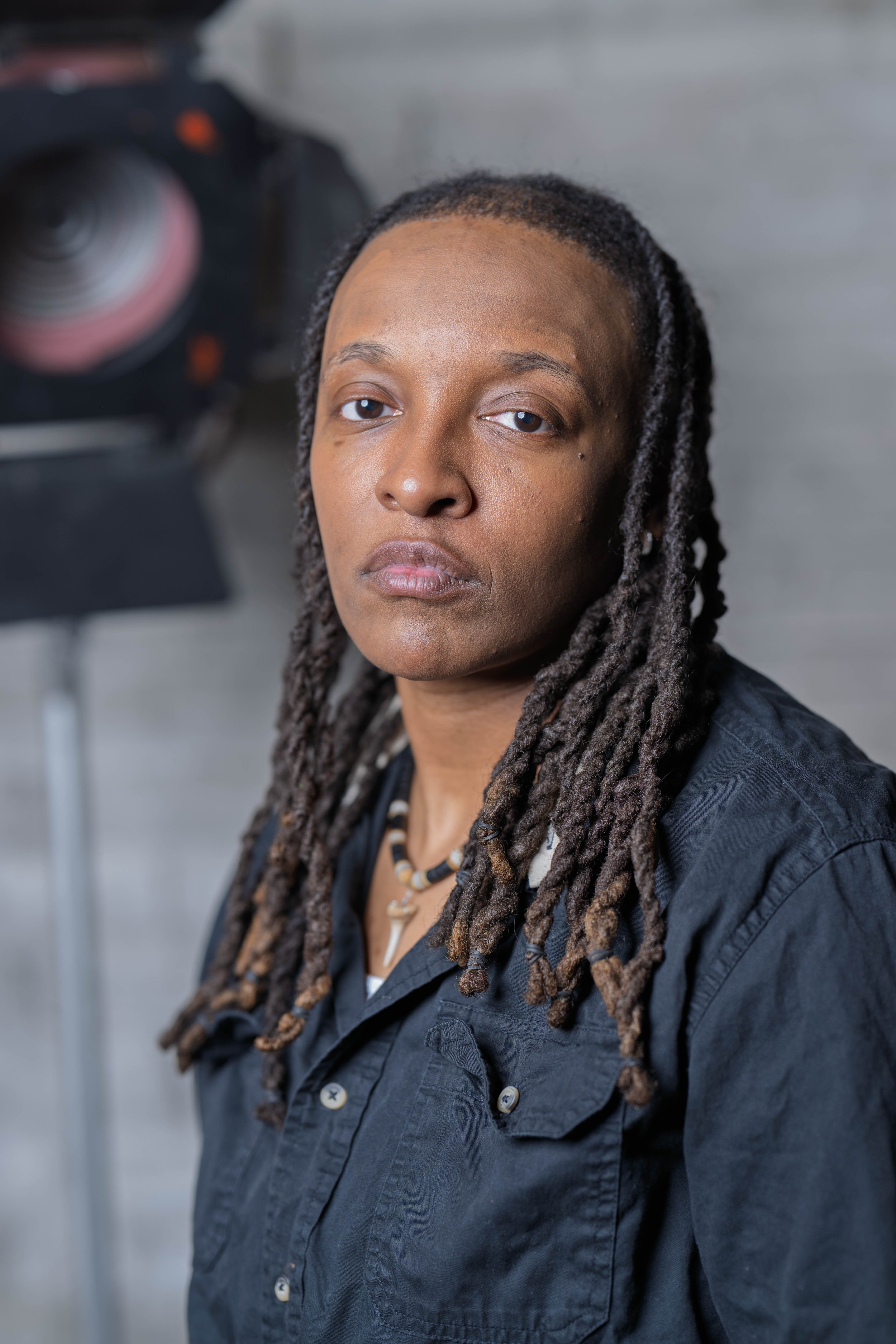 Portrait of a woman with long dreadlocks, wearing a dark shirt, standing in front of grey wooden background, with a spotlight visible in the background.