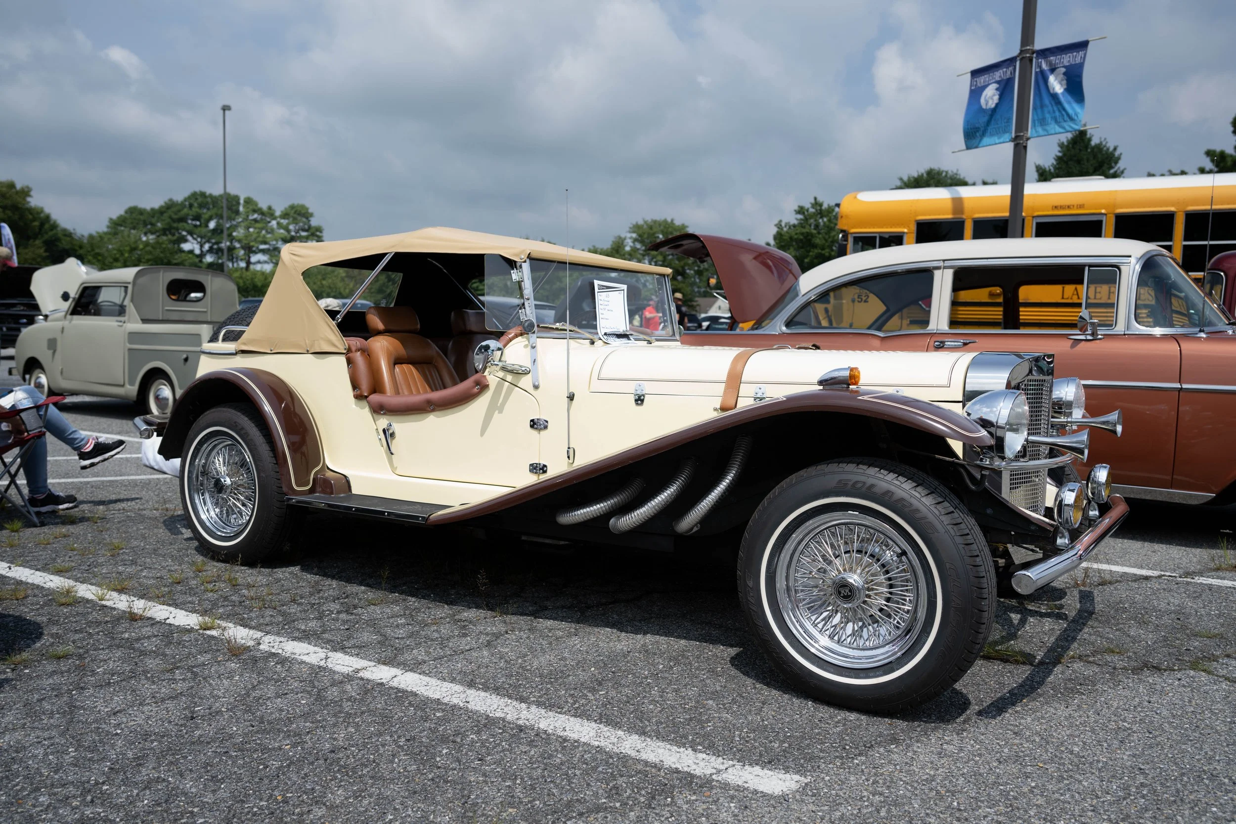 A vintage cream and brown open-top car with leather seats parked among other classic cars at a car show on a cloudy day.