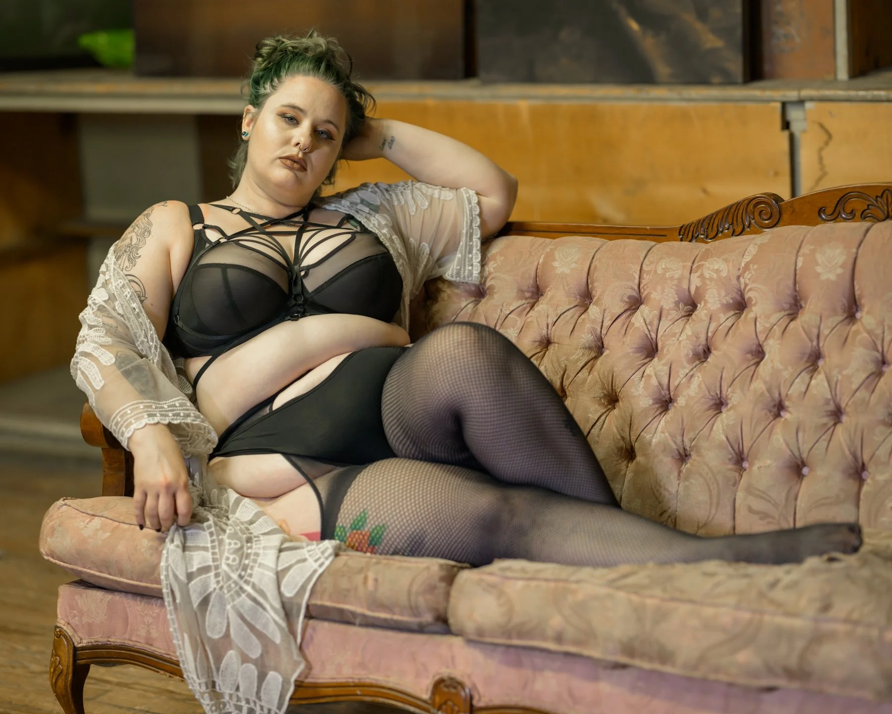 A woman with green hair and tattoos reclining on an antique pink sofa, wearing black lingerie, fishnet stockings, and a sheer cardigan.