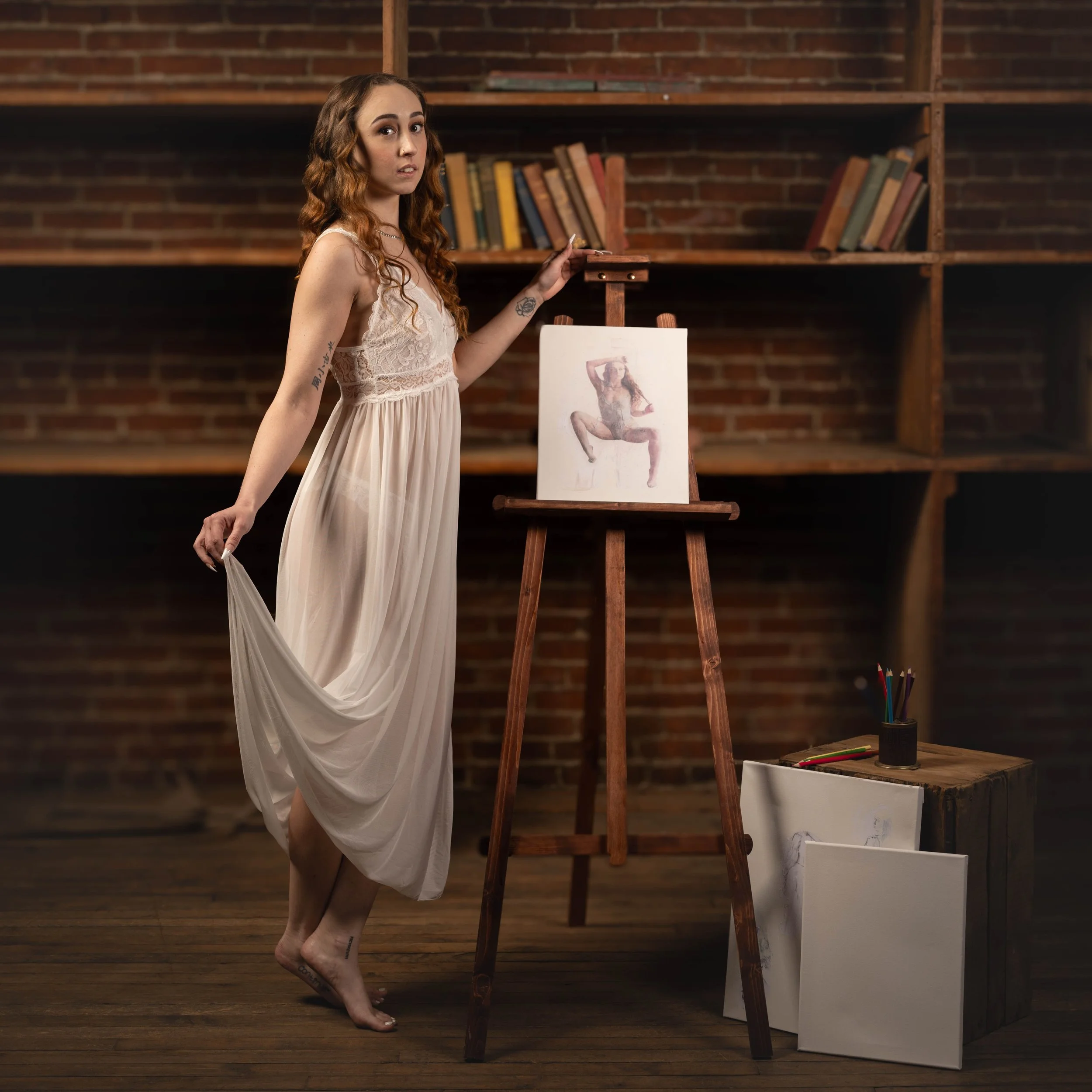 A woman in a flowing, light-colored dress standing barefoot in an art studio with brick walls. She is holding the hem of her dress with one hand and has her other hand on a wooden easel displaying a painting of a woman in a similar pose. There are ar