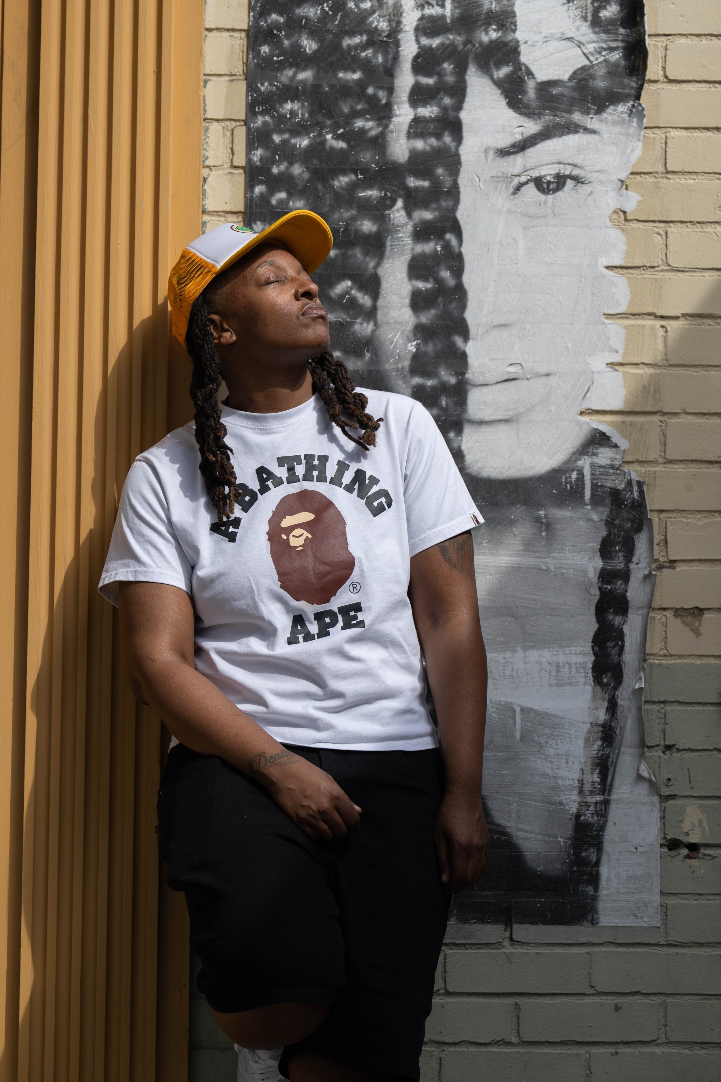 Person leaning against a wall with a portrait painting behind them. The person is wearing a white T-shirt with the 'A Bathing Ape' logo, a yellow and white trucker hat, and has long dreadlocks.