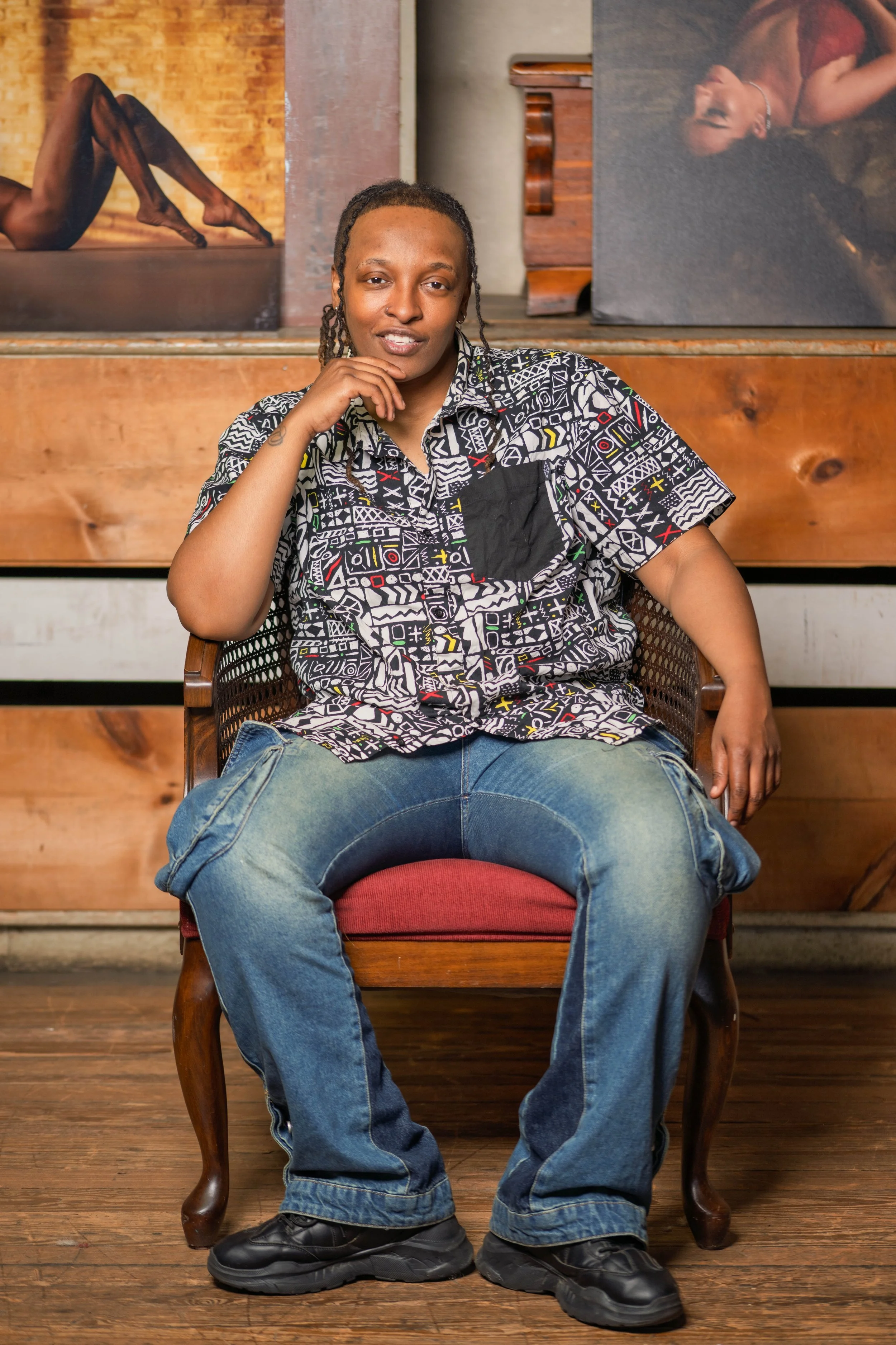 A person sitting on a wooden chair with a red cushion, wearing a patterned shirt and jeans, in an art gallery.