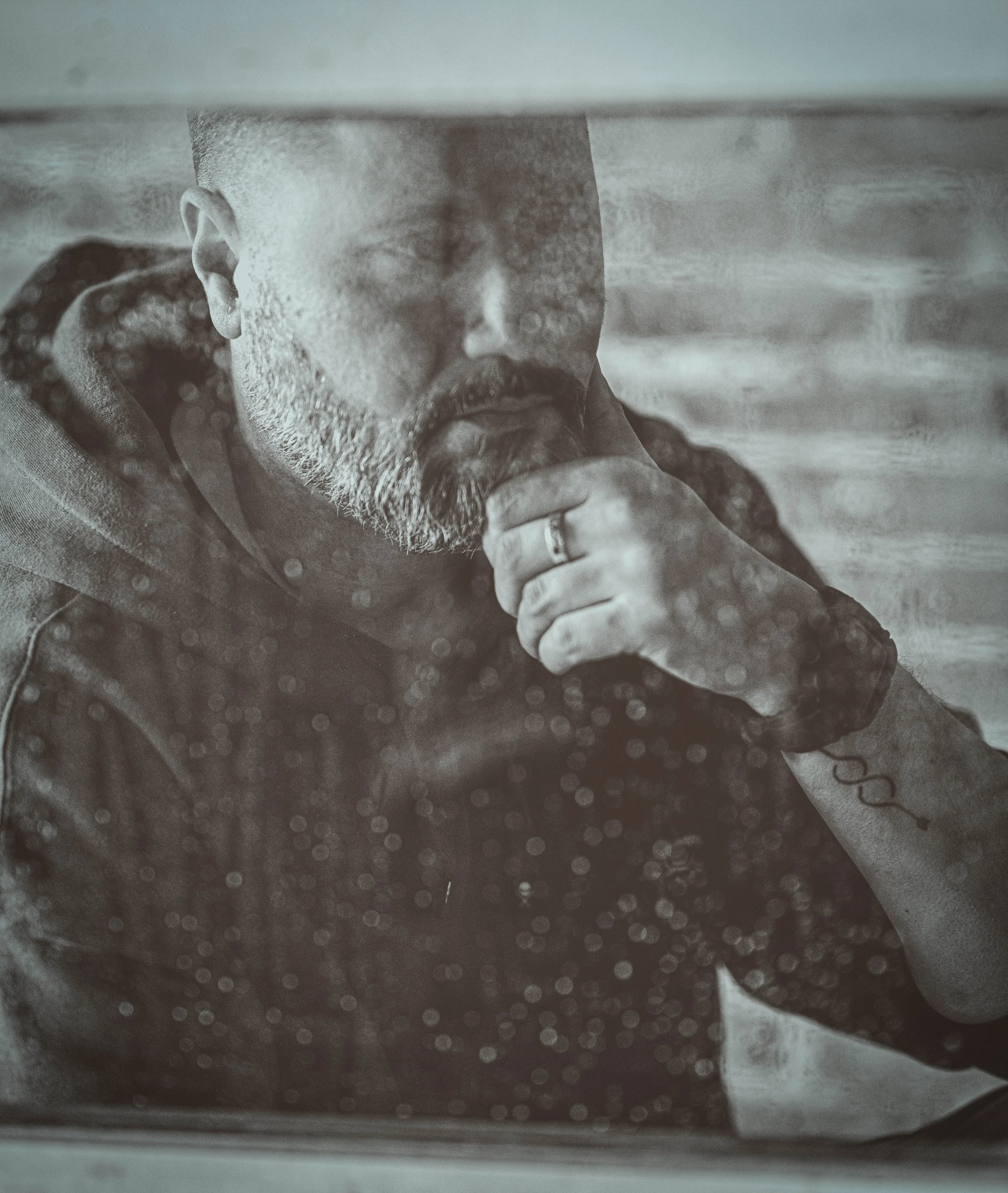 A man with a beard and short hair, wearing a hoodie, has his hand on his chin and is looking thoughtfully through a rain-spattered window.