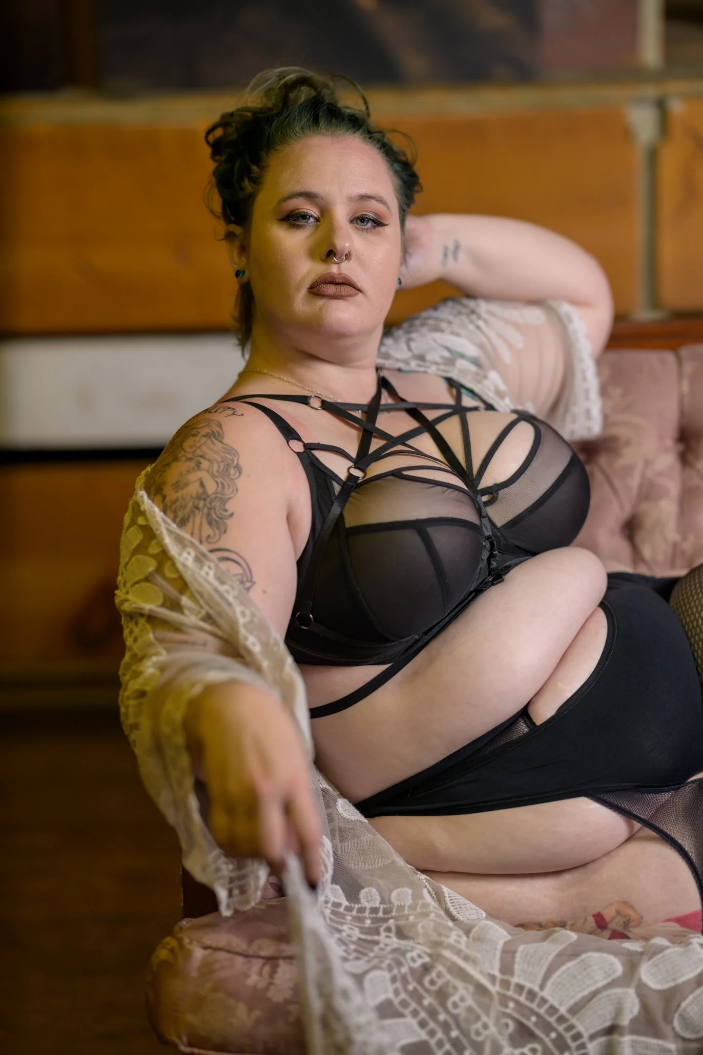 A woman with tattoos and piercings sitting on a pink velvet sofa, wearing black lingerie, a lace cover-up, and thigh-high stockings, with her arm resting behind her head and looking at the camera.
