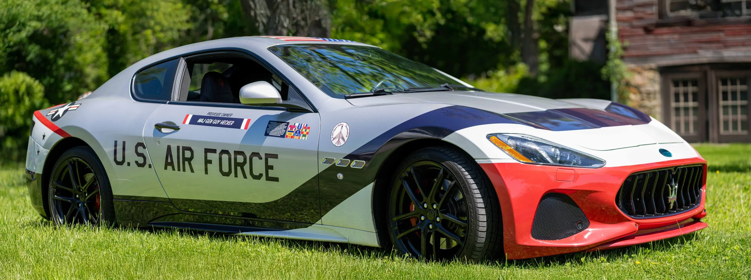 A sports car with U.S. Air Force markings parked on a grassy area, with trees and a building in the background.