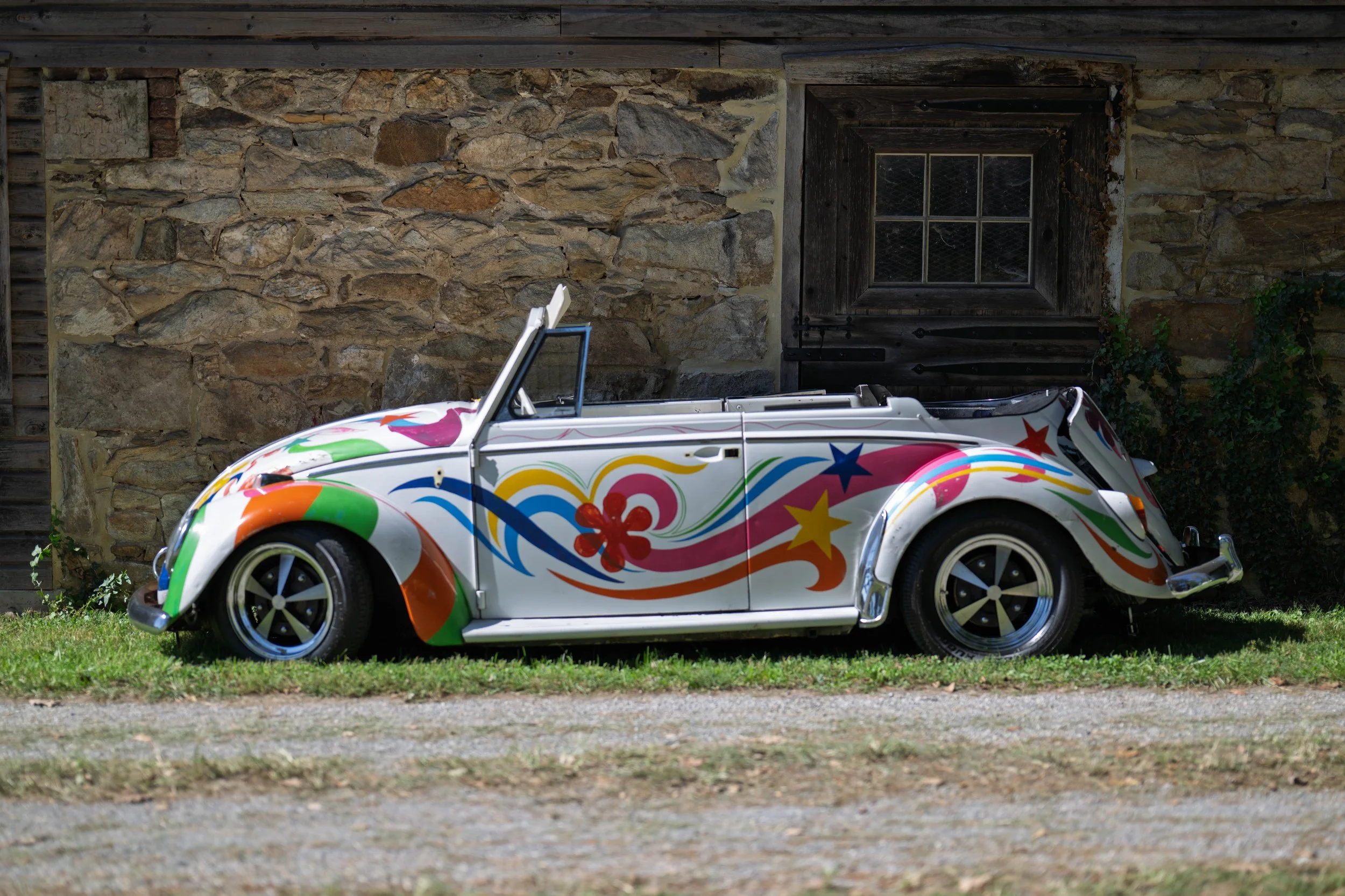 Colorful vintage convertible car with floral and star designs parked on grass beside a stone building with a wooden window.